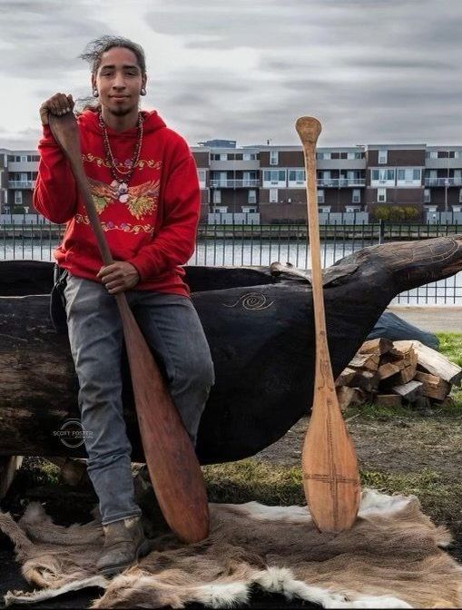 Man in red hoodie leans against a canoe, holding a paddle. Second paddle beside him. Outdoors, by water.