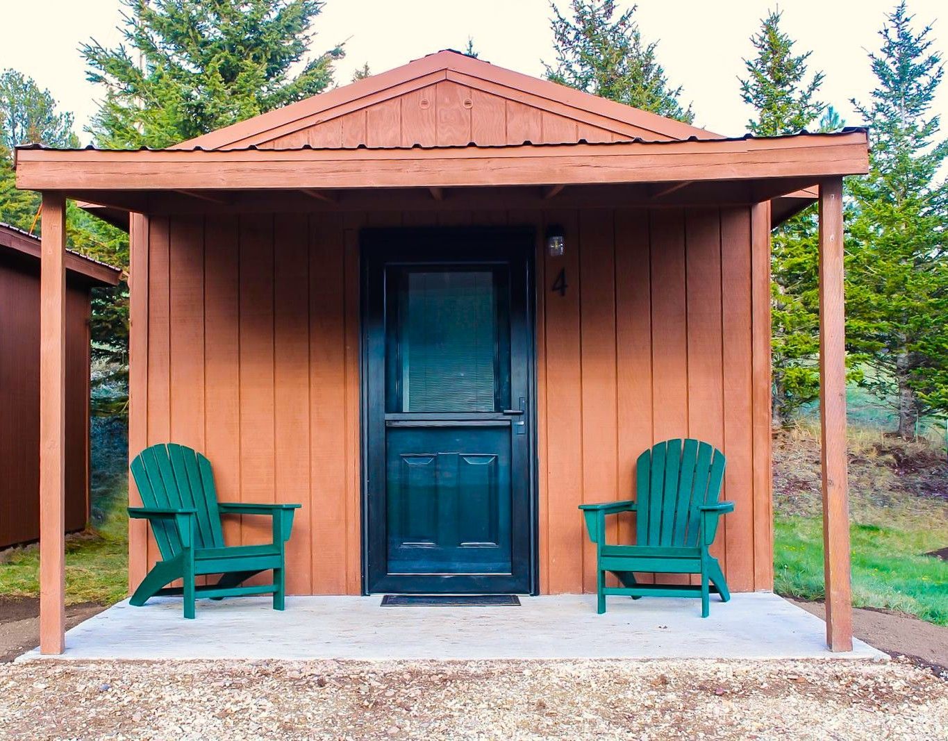 Small wooden cabin porch with a teal door and two green chairs, surrounded by trees.