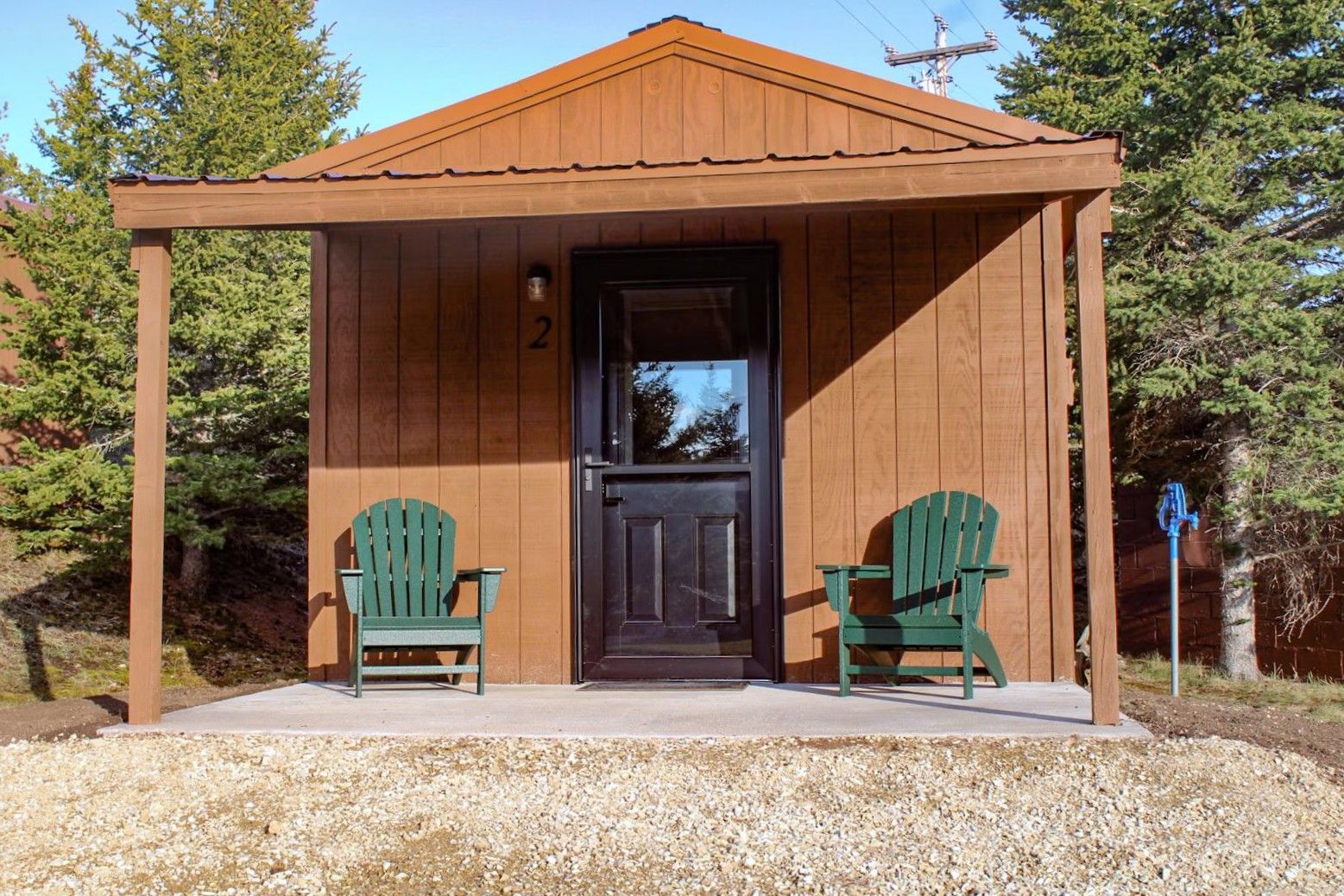 Small brown cabin with a front porch, black door, and two green chairs in a wooded setting