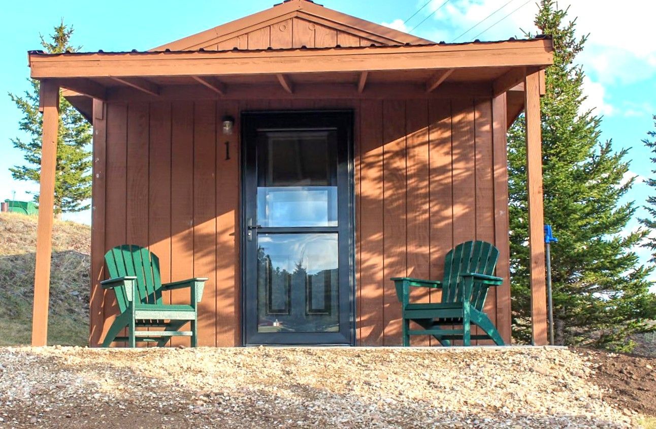 Small brown cabin with a black door and two green chairs on the porch, surrounded by trees.