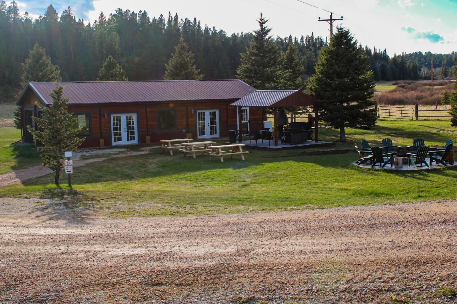 Rustic cabin with picnic tables in a grassy field beside a gravel road and forest