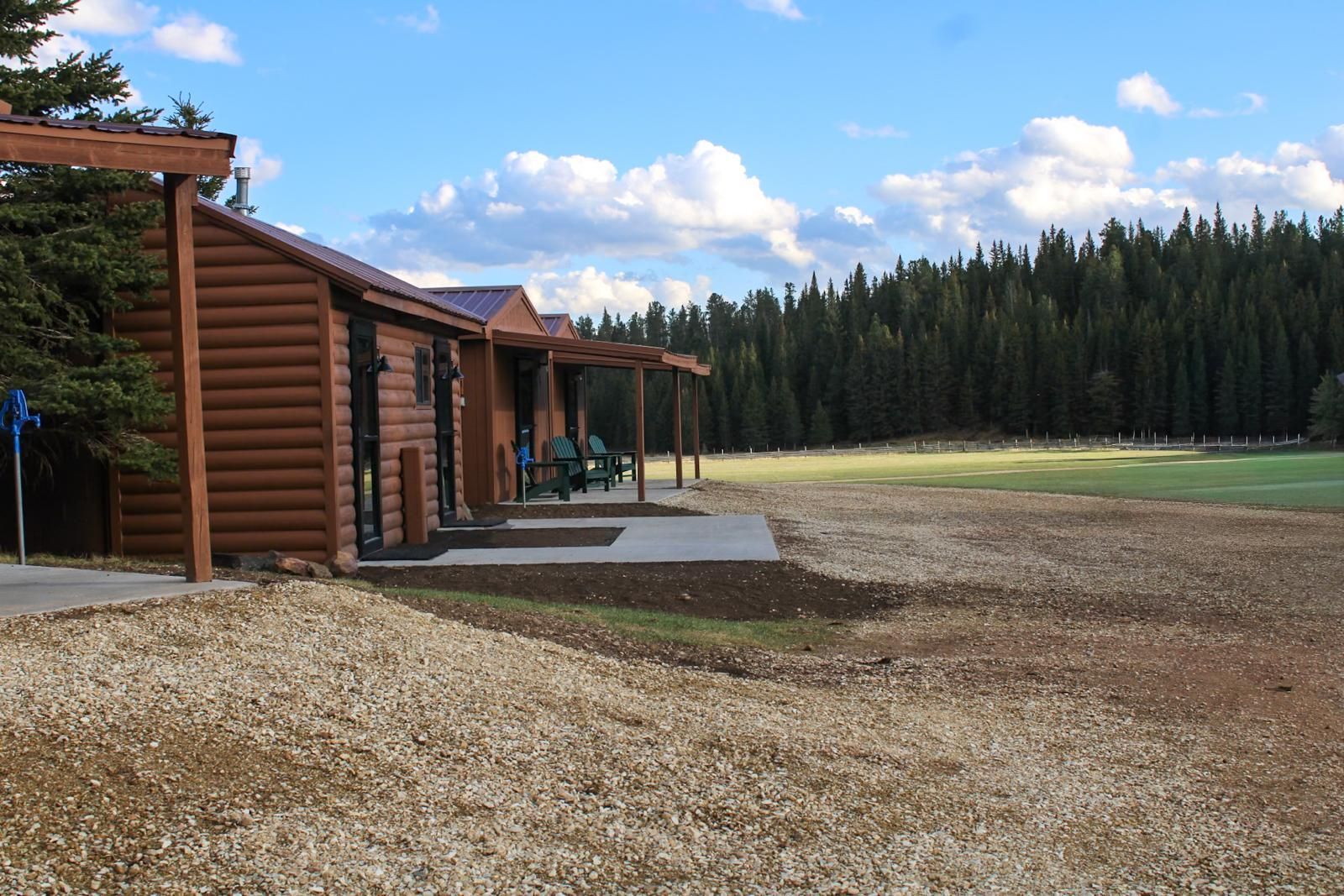 Log cabins beside a gravel path and open field, with pine trees and a blue sky in the background