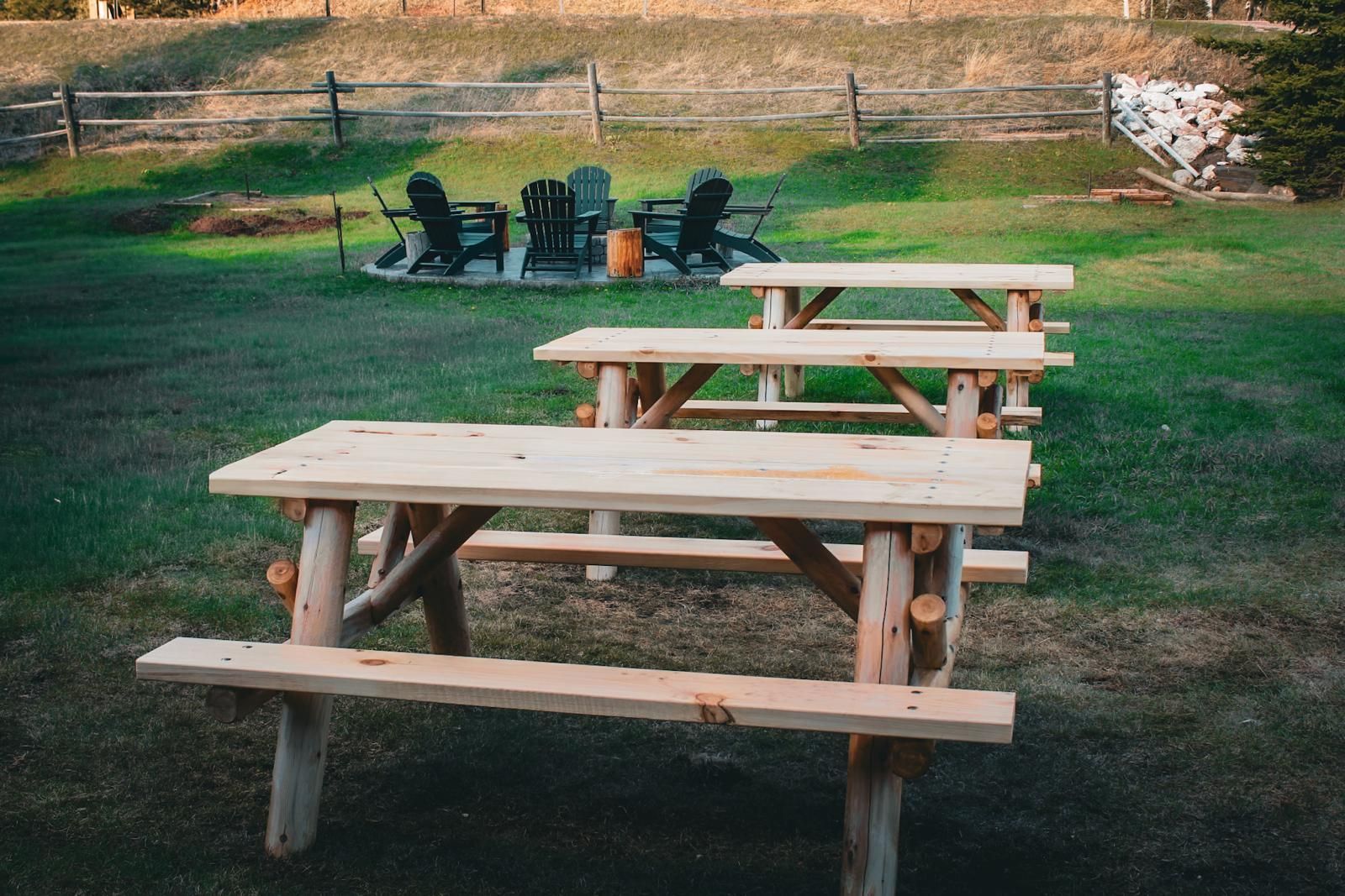Wooden picnic tables on a grassy field with a fire pit and chairs in the background
