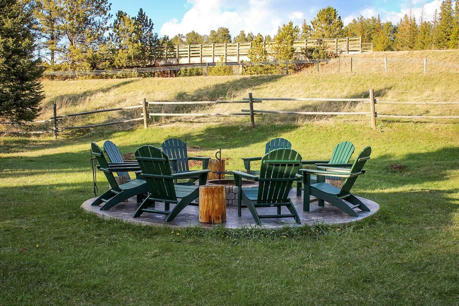 Outdoor fire pit with green chairs on a grassy lawn beside a wooded hillside
