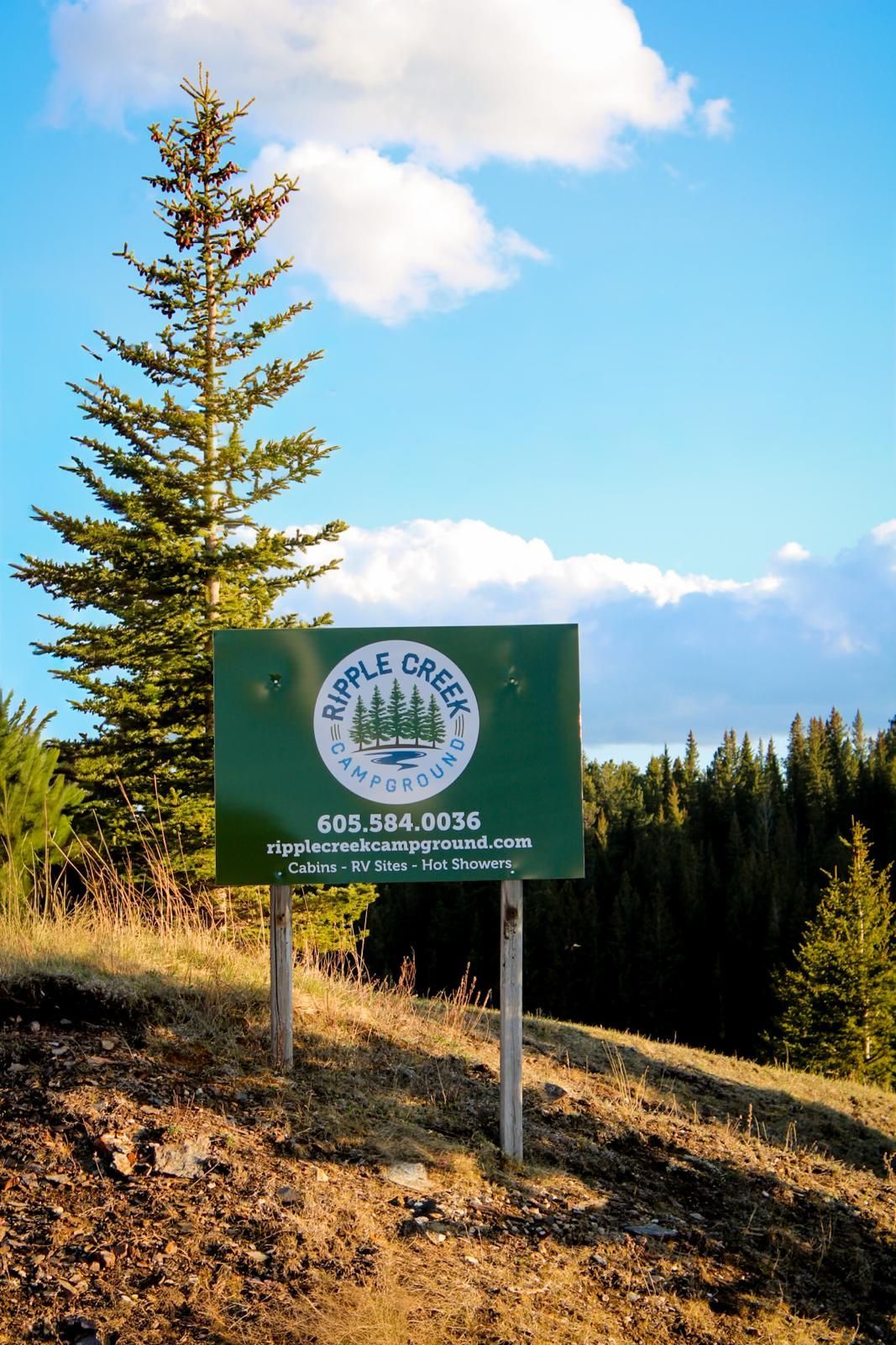 Green roadside sign beside a dirt hill and pine trees under a blue sky with clouds