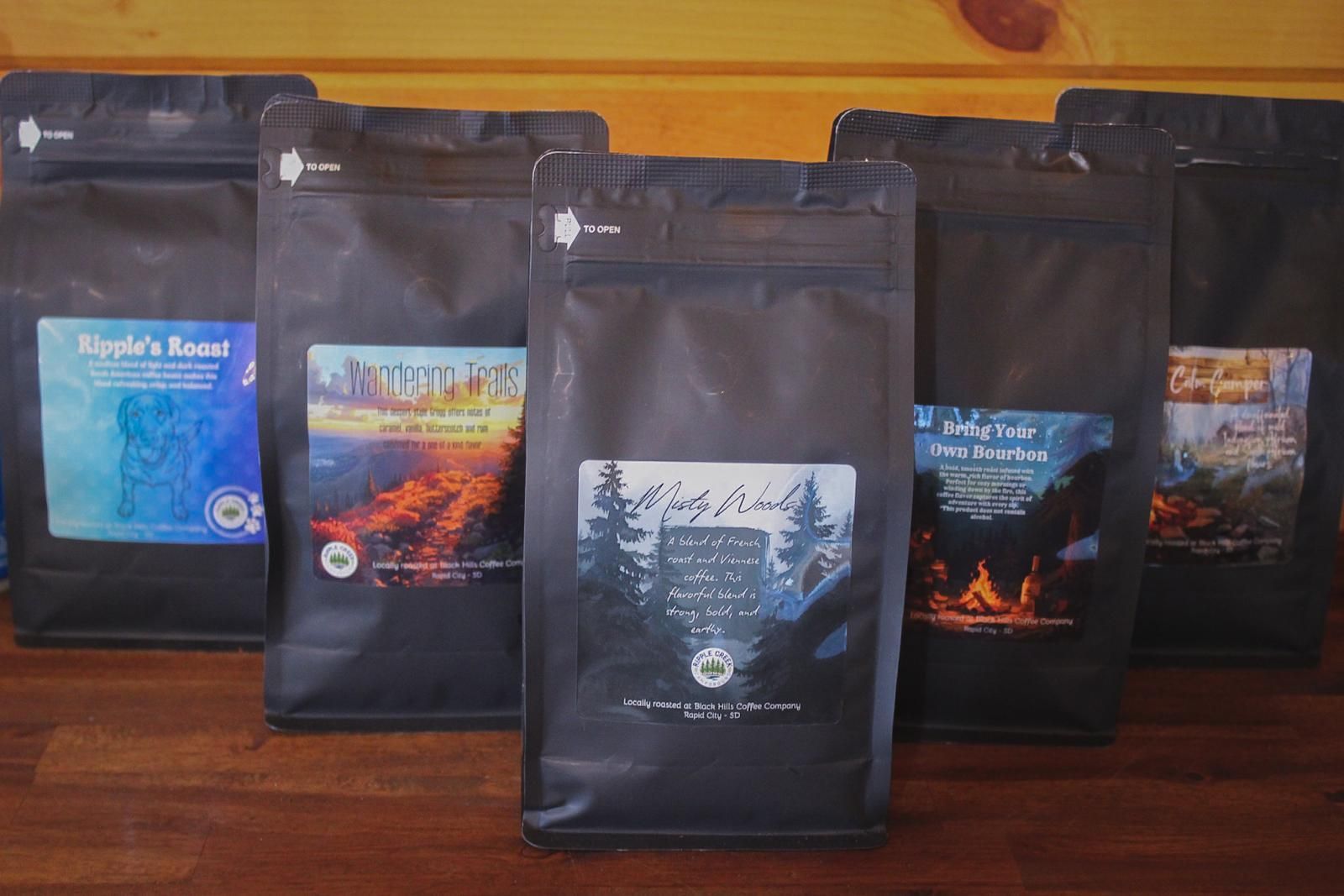 Five coffee bags in assorted dark packaging lined up on a wooden table against a yellow wall.