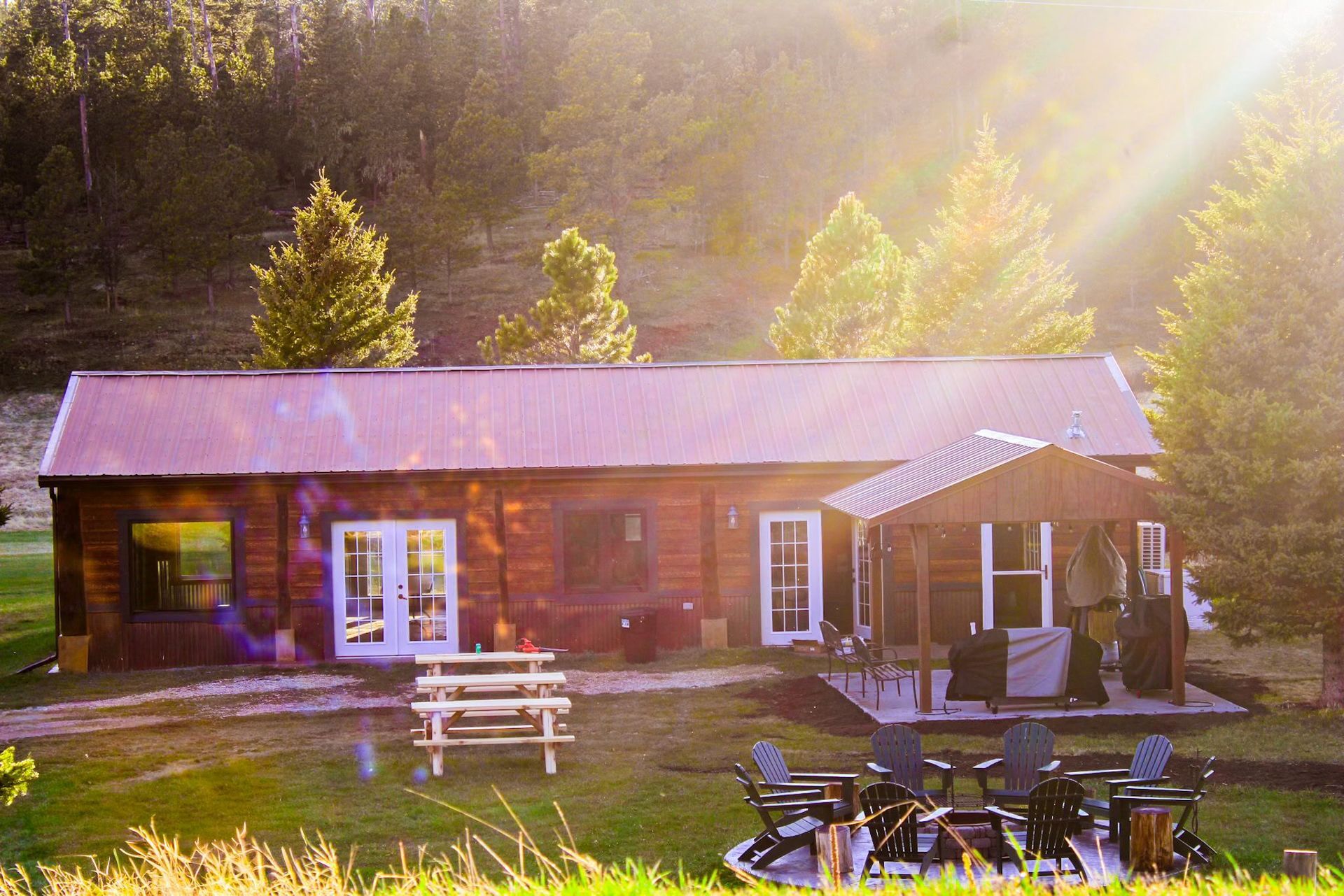 Sunlit rustic cabin with porch, picnic table, and outdoor chairs in a wooded meadow