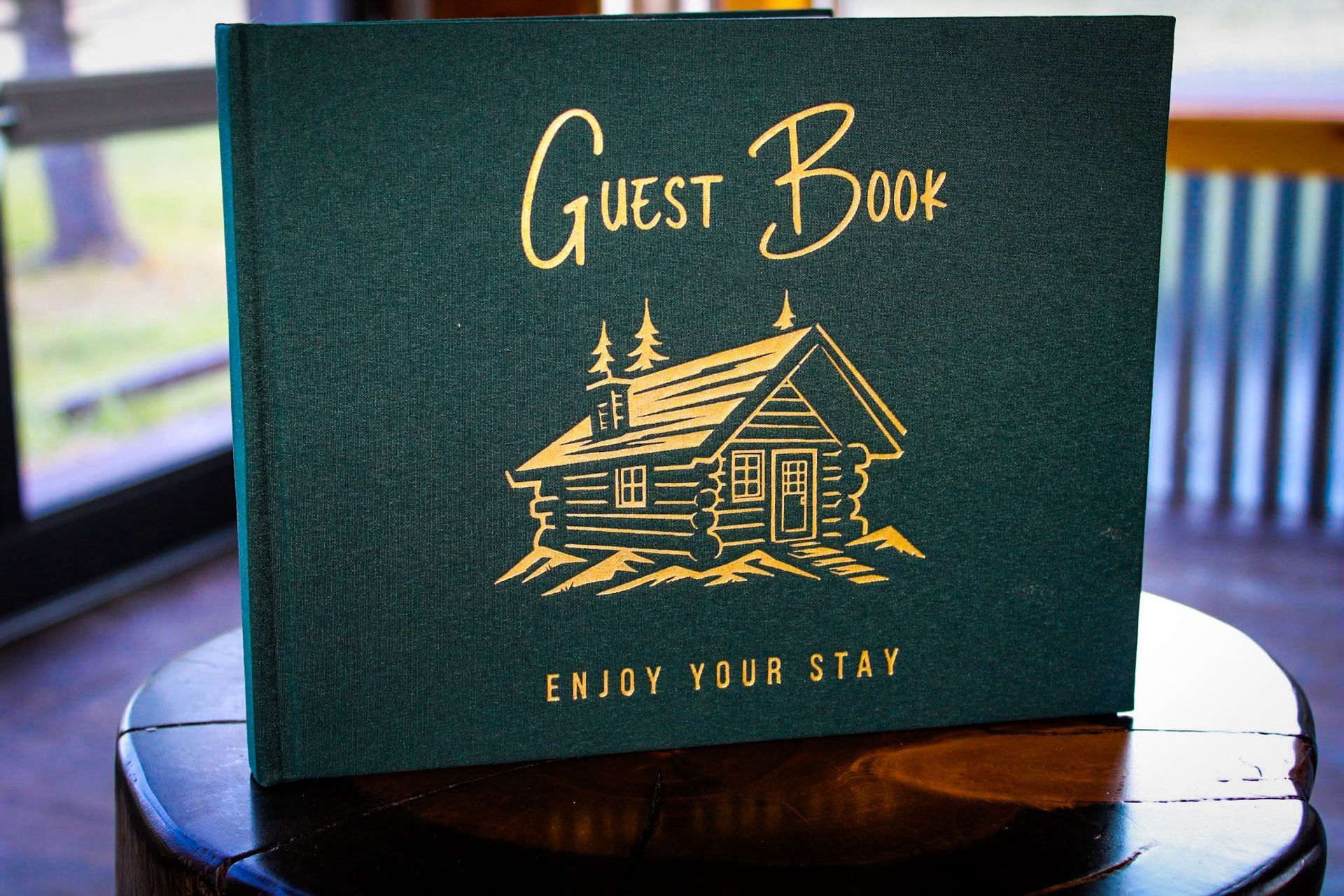 Green guest book with gold cabin illustration and “Enjoy Your Stay” text on a round table