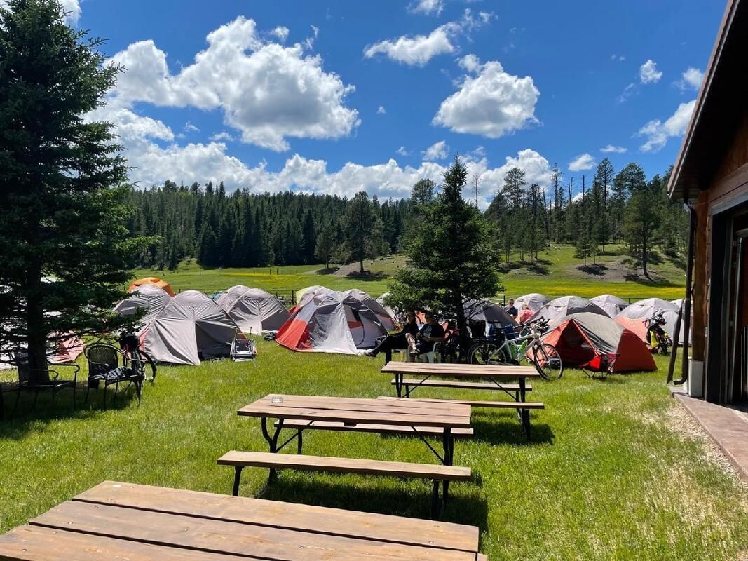 Camping tents on a sunny grassy field beside picnic tables and a lodge under a blue sky
