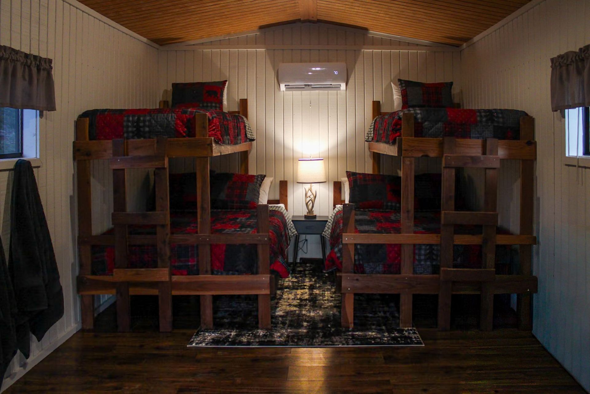 Dorm-style room with two bunk beds, patterned blankets, a window at the back, and wooden ceiling and floor