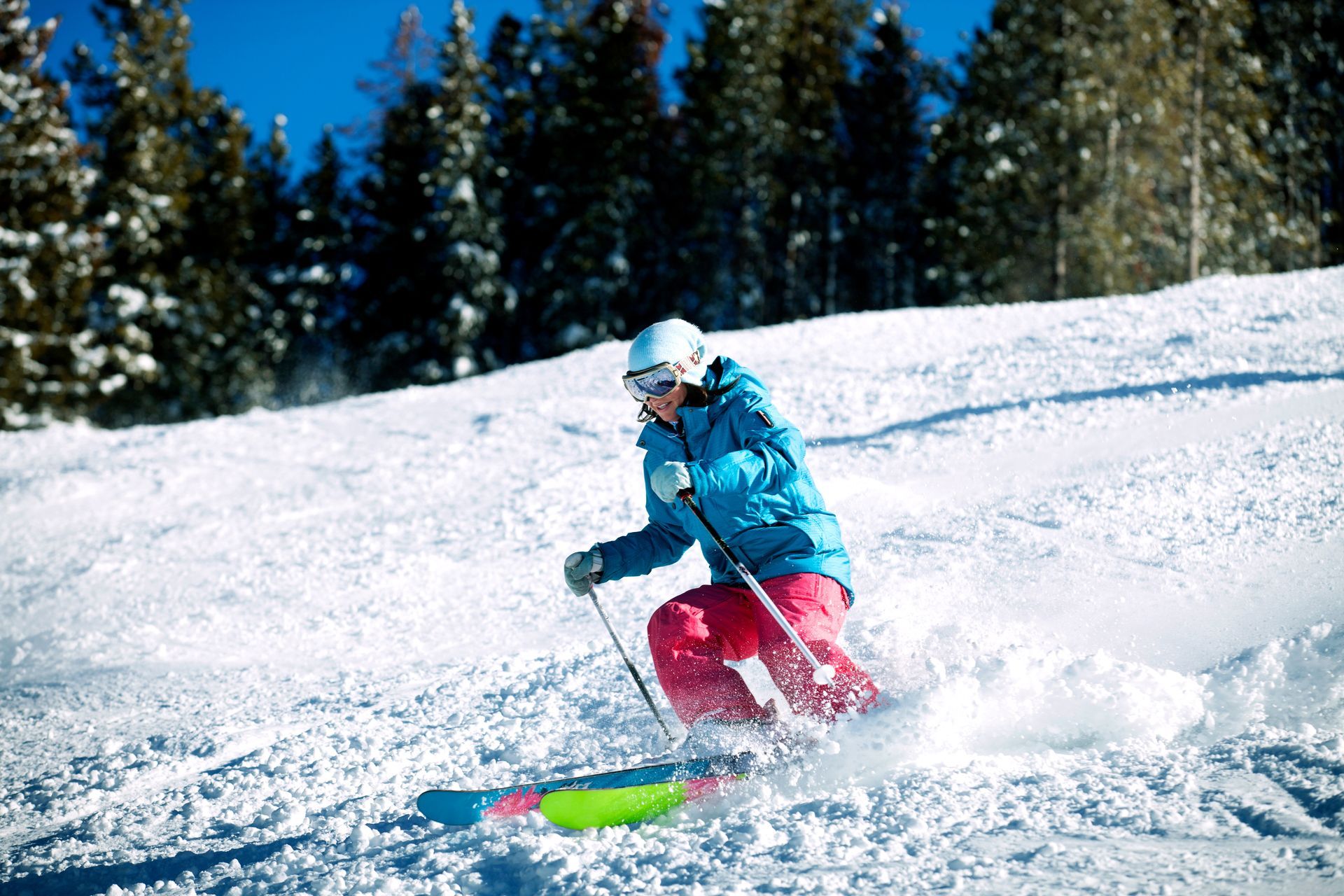 Skier in blue jacket and red pants carving through snow on a sunny slope with evergreen trees behind.