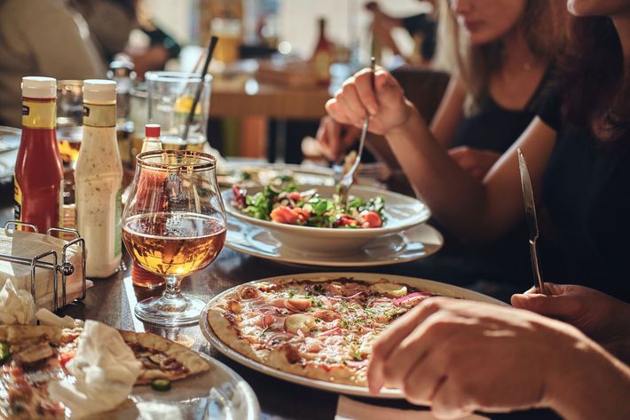 Friends dining at a table with pizza, salad, and drinks in a warm restaurant setting