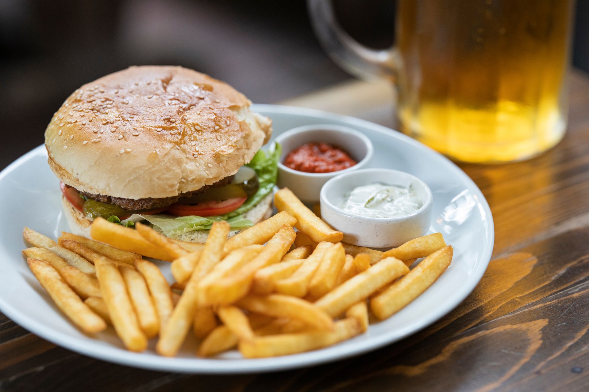 Burger and fries on a white plate with ketchup and dipping sauce, beside a glass of beer on a table