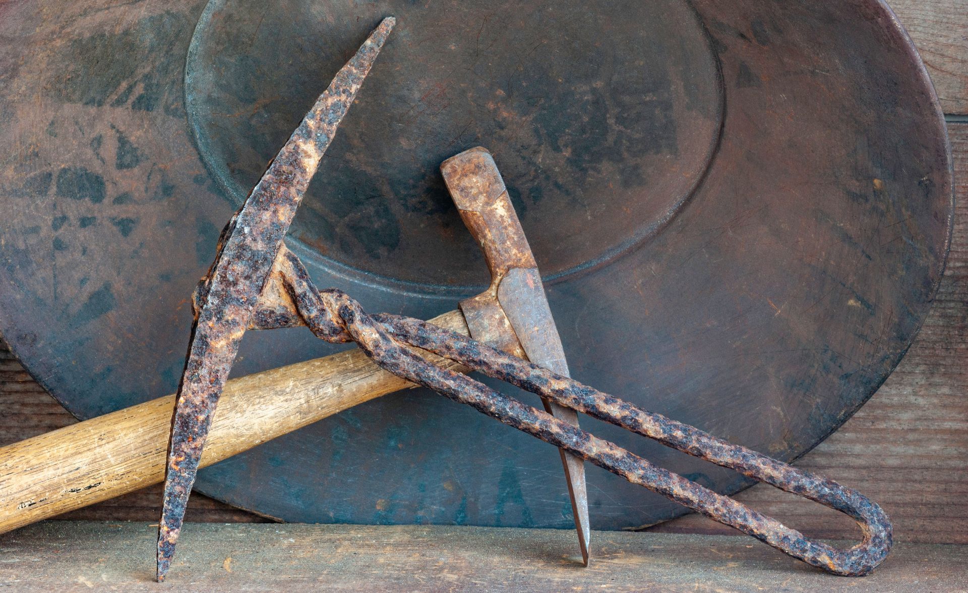 Rusty pickaxe and hammer crossed over a large blacksmith’s anvil on a wooden surface.