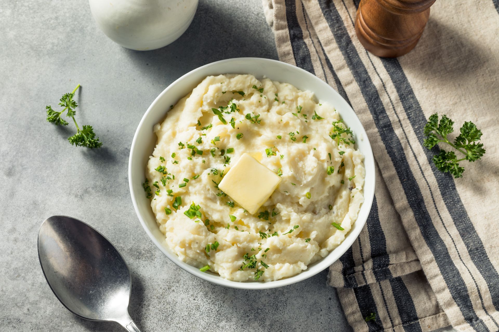 Bowl of creamy mashed potatoes topped with butter and herbs on a gray table, with spoon and pepper mill nearby