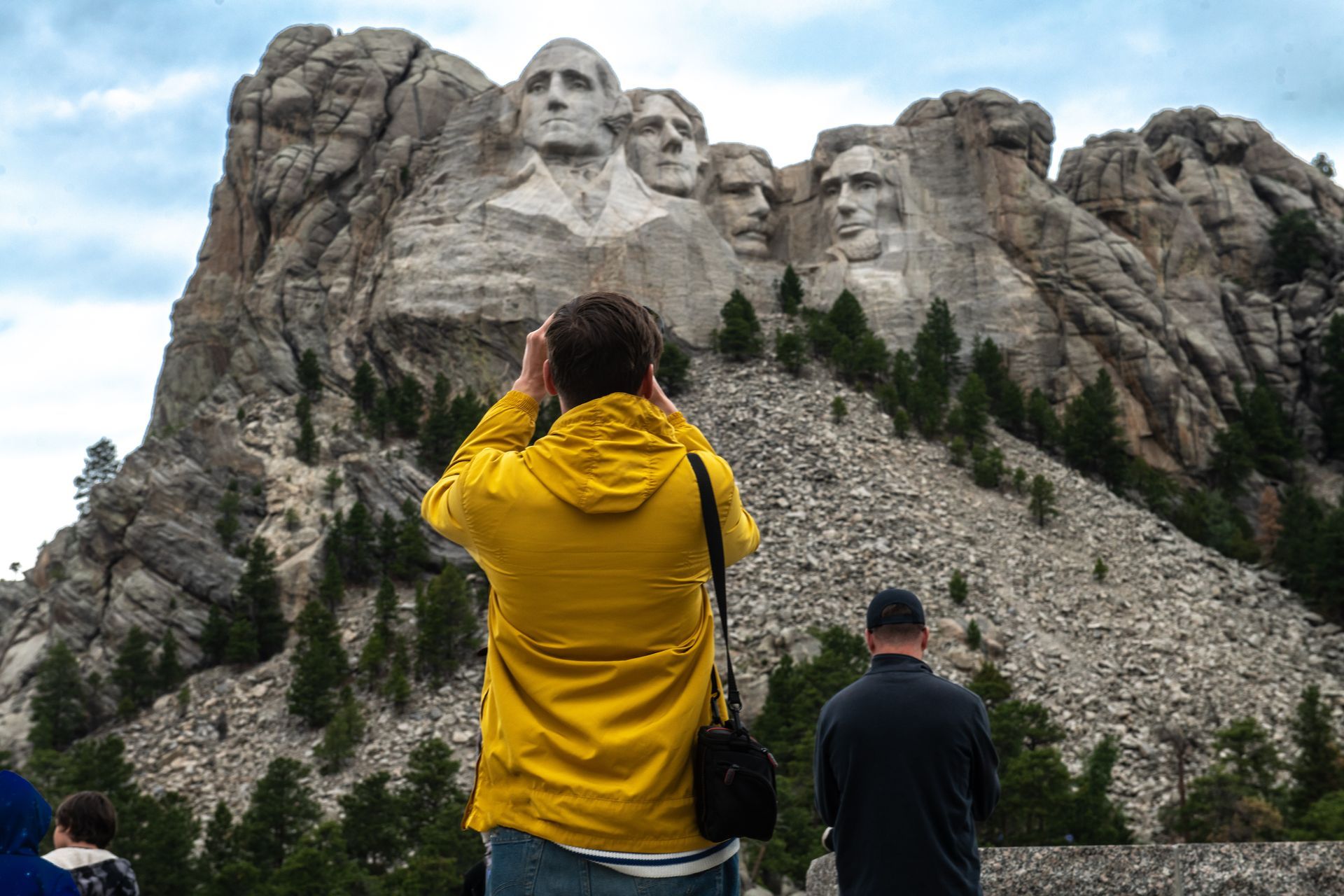 Visitors photograph Mount Rushmore under cloudy skies, one in a bright yellow jacket.