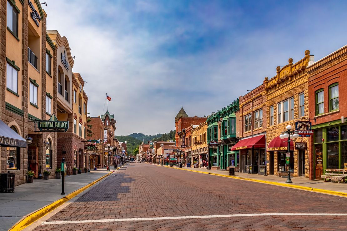Historic small-town main street with colorful brick storefronts, cobblestone road, and mountains in the distance