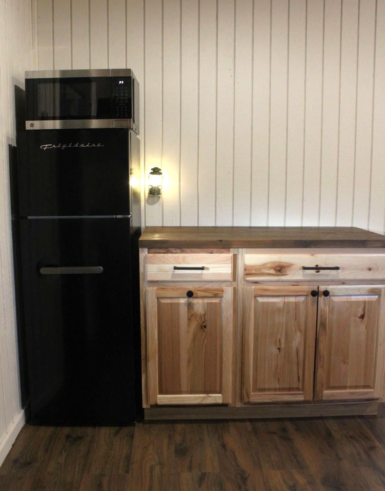 Black refrigerator beside light wood cabinets in a rustic kitchen with vertical white wall paneling.