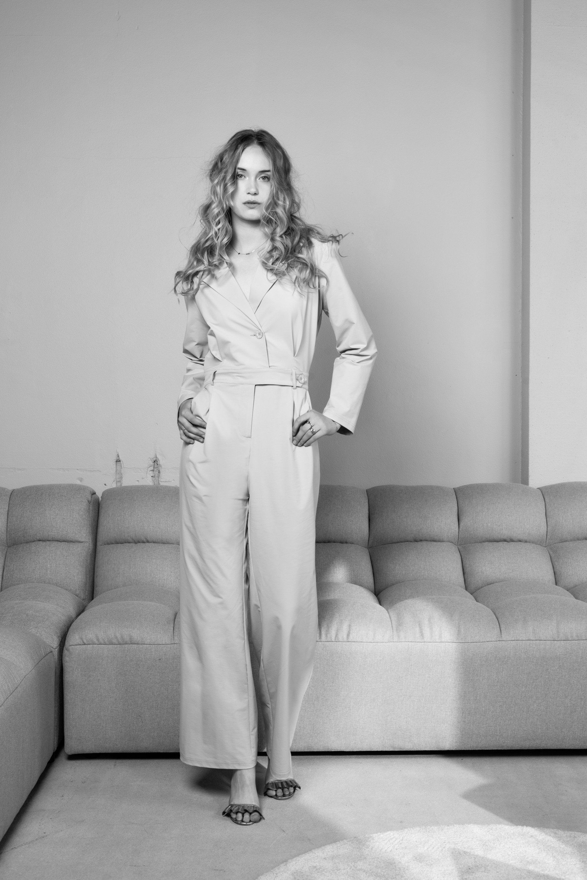 Vrouw staat in een woonkamer naast een bank, gekleed in een lichtgekleurde outfit.