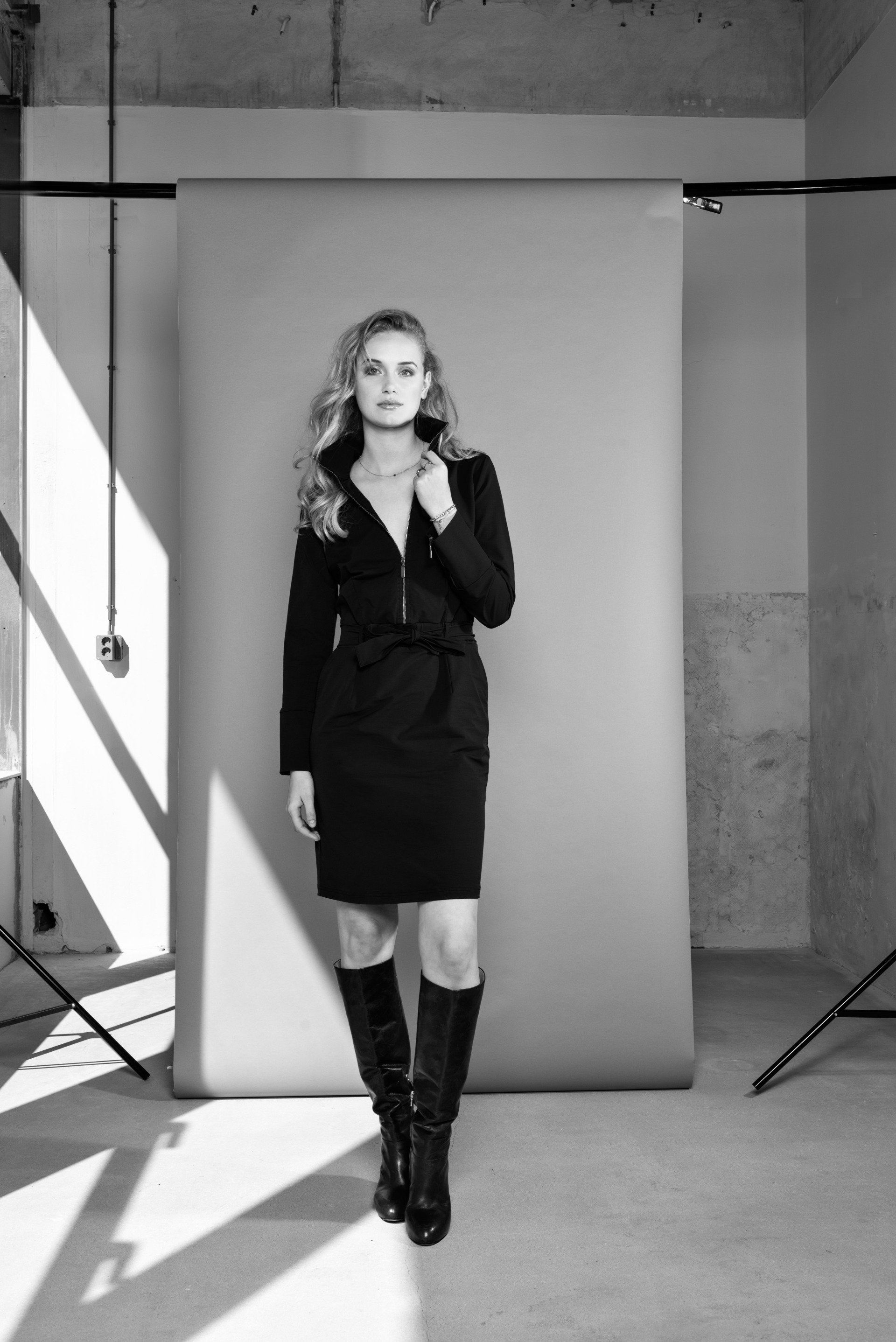 Vrouw in een zwarte jurk en laarzen poseert in een studio met een grijze achtergrond en statieven voor de verlichting.