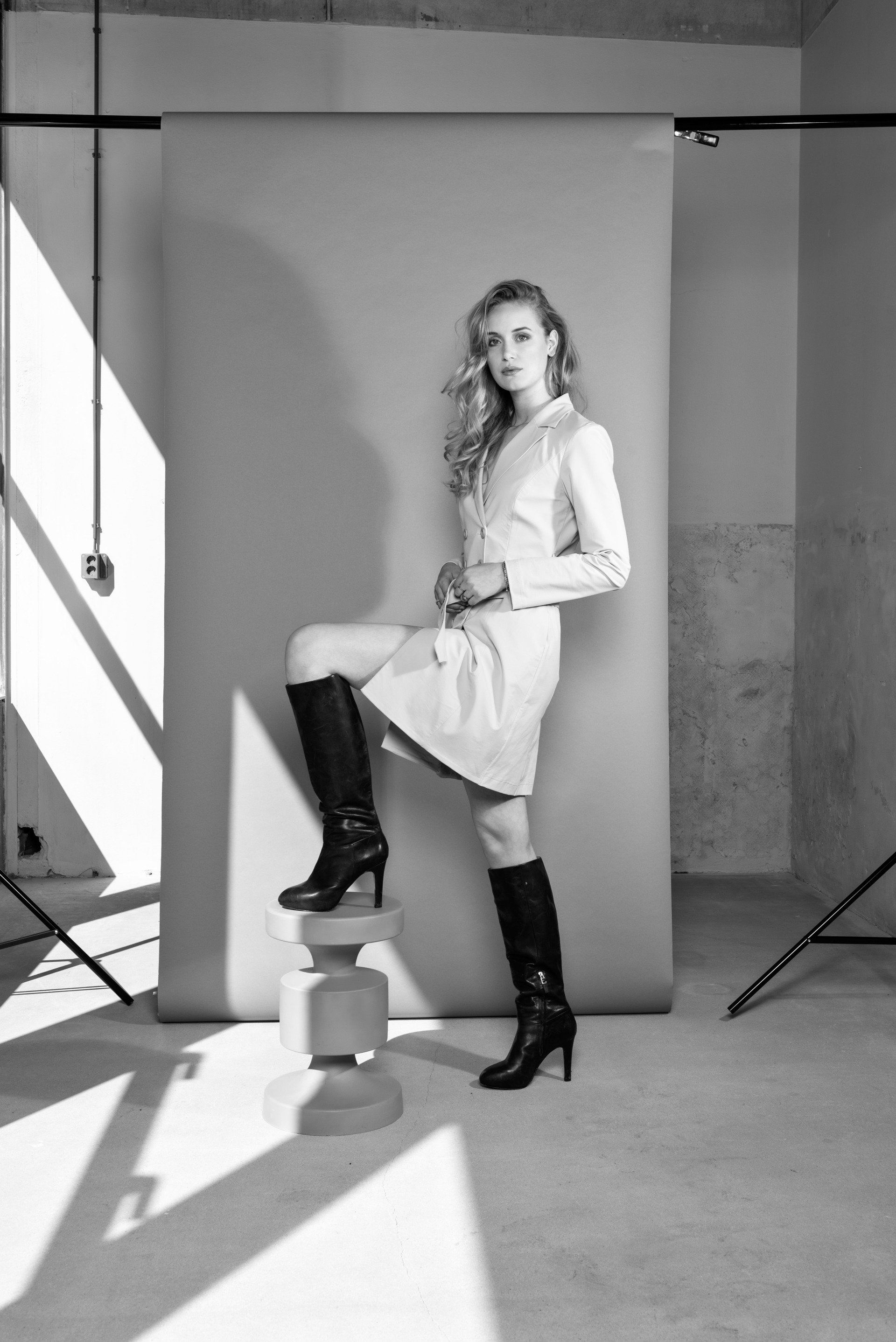 Vrouw poseert in een studio, met één voet op een krukje, gekleed in een lichte jas en zwarte laarzen.