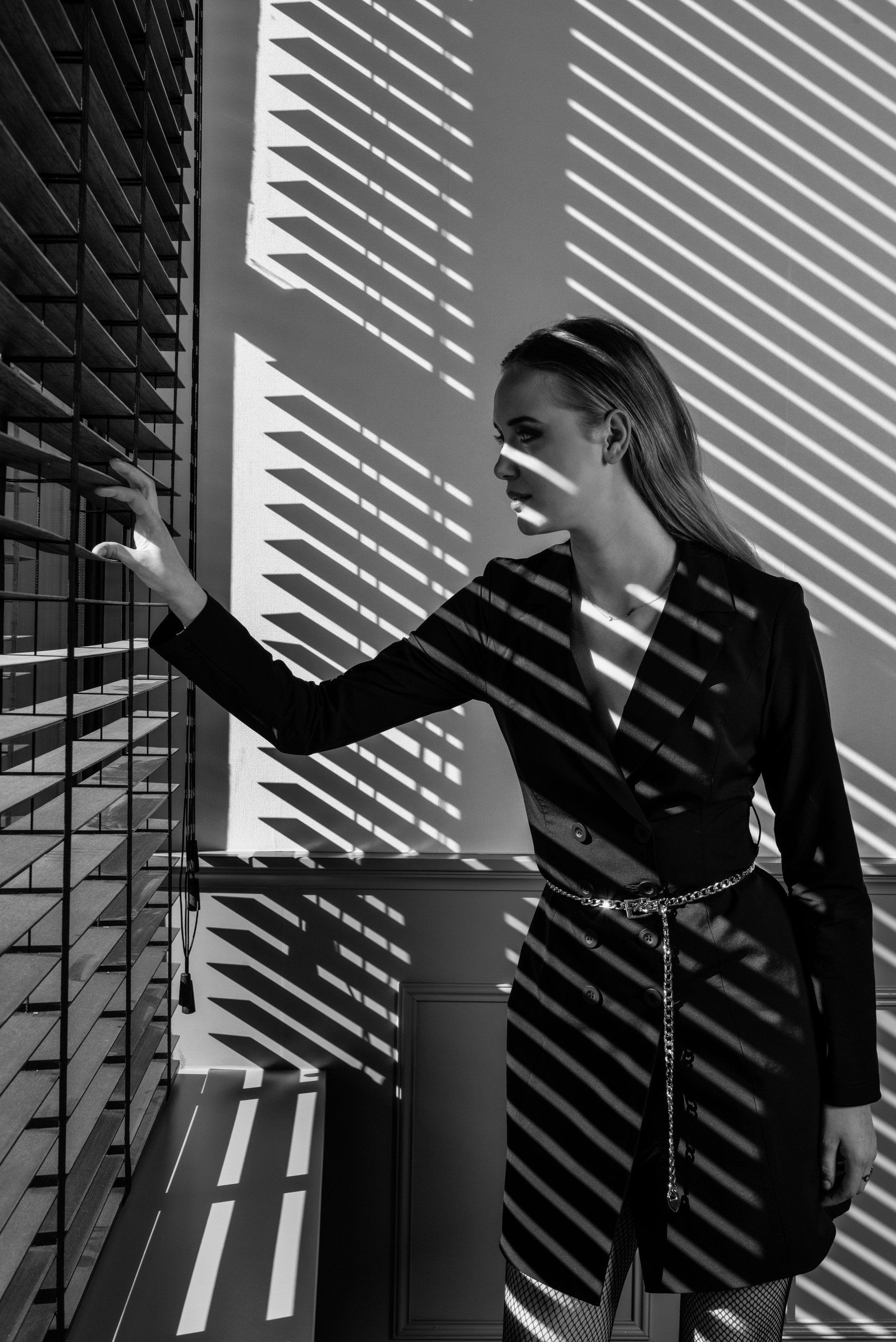 Vrouw in een zwarte jurk raakt een muur aan met gestreepte licht- en schaduwpatronen.