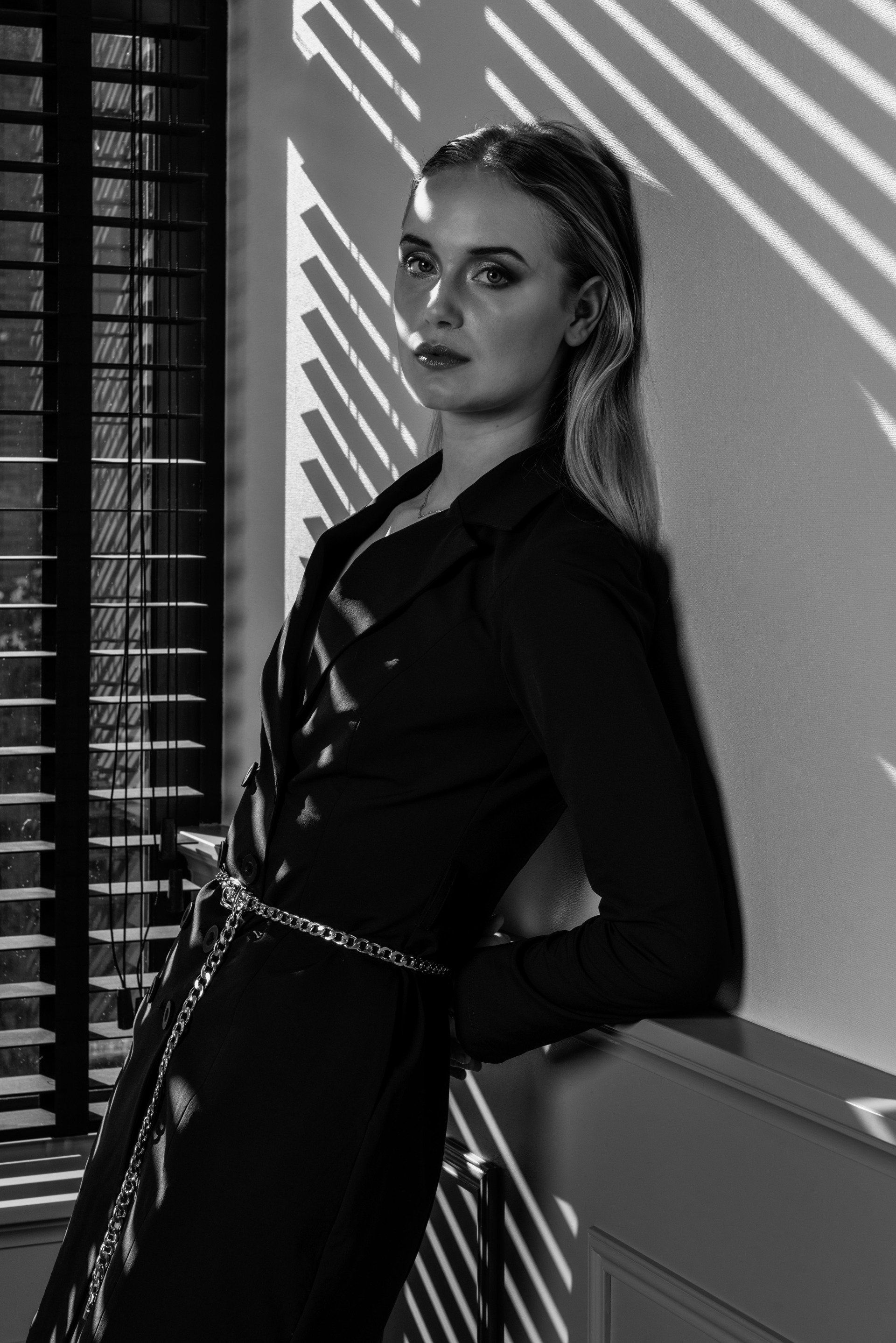 Zwart-wit portret van een vrouw die poseert naast een muur met gestreepte lichtpatronen.