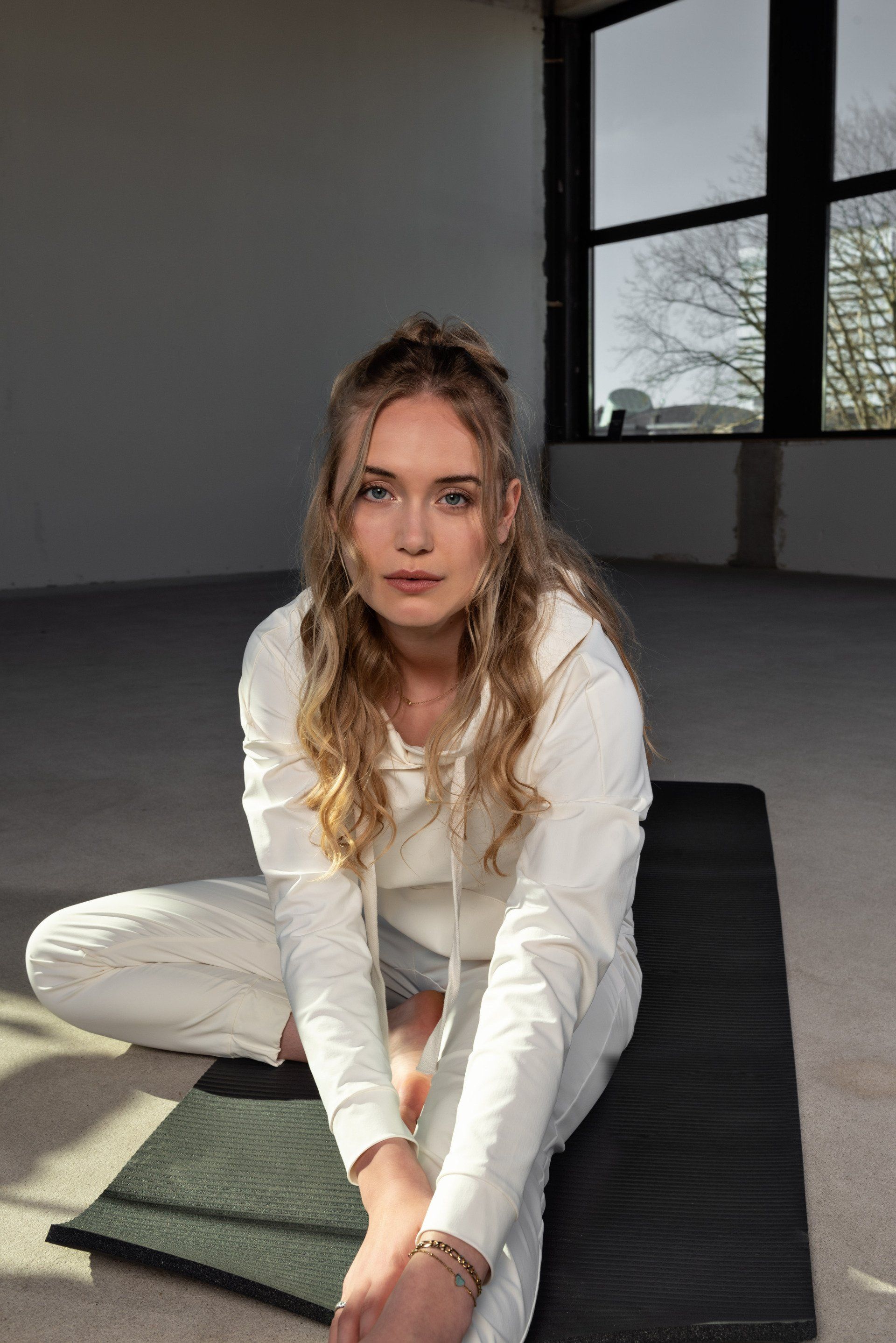 Vrouw in witte kleding zit op een mat in een lichte, lege studio vlakbij grote ramen.