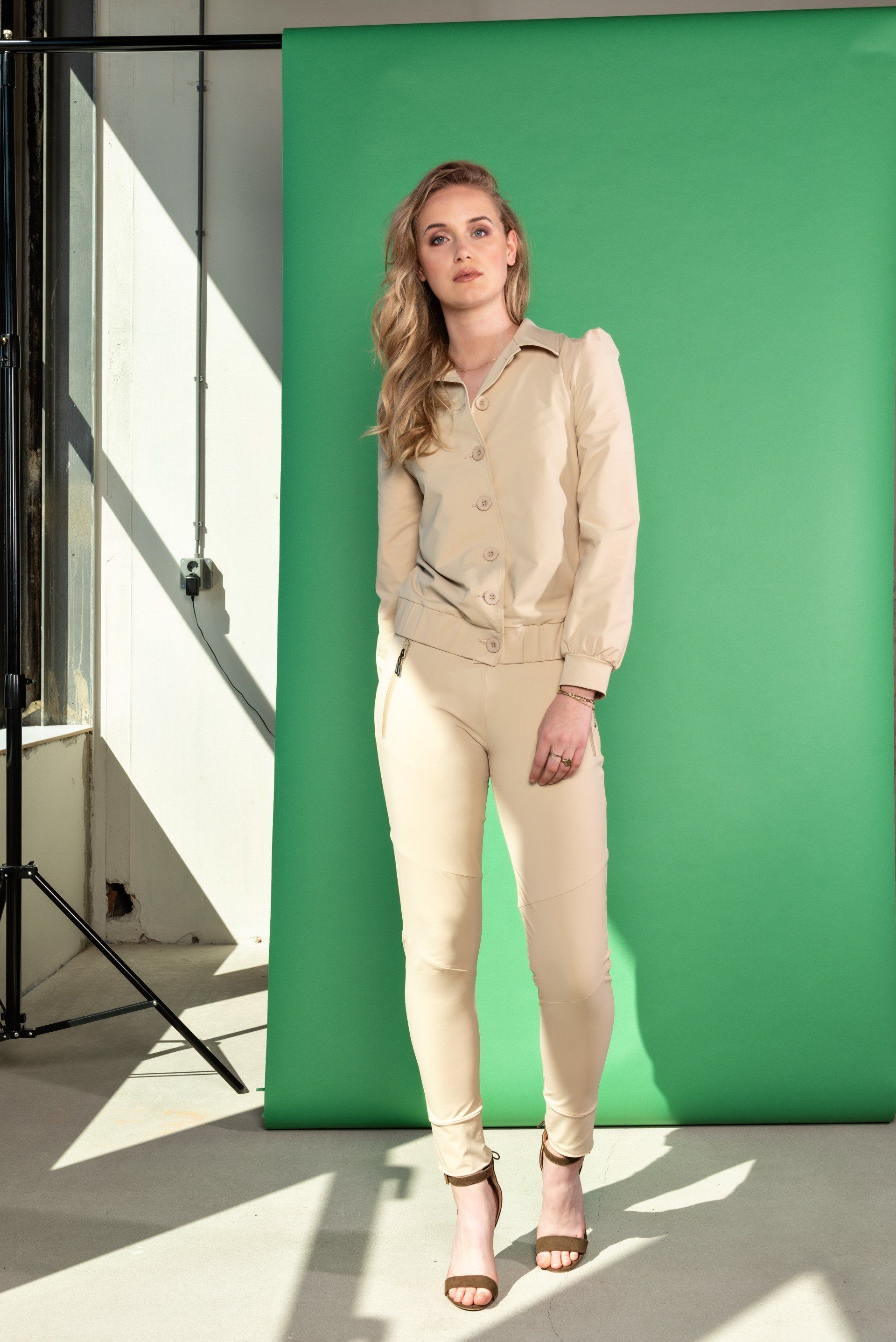 Vrouw staat in een studio met een groene achtergrond, gekleed in een beige outfit en hakken.