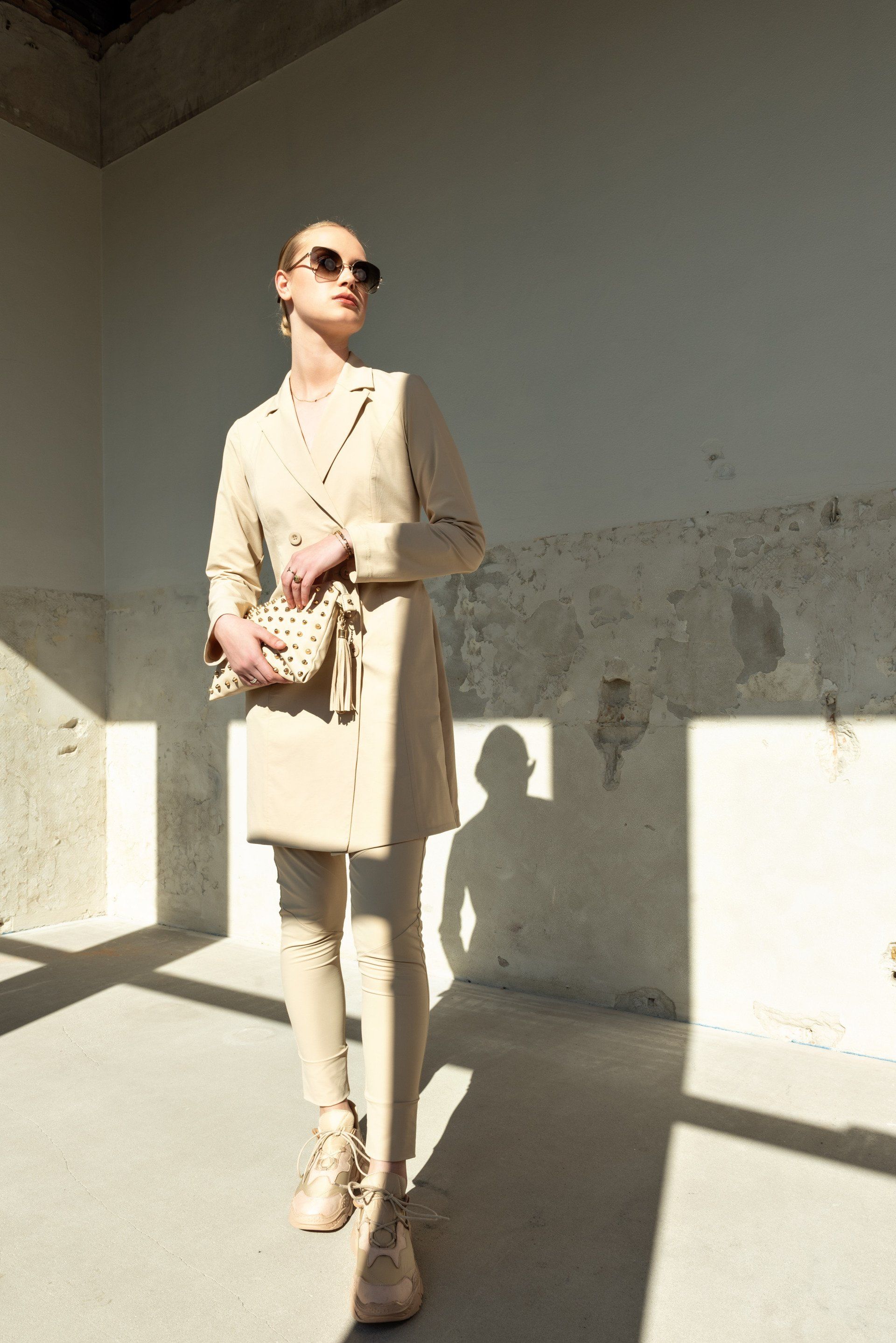 Vrouw in beige jas en zonnebril staat in een zonovergoten galerij, met een handtas met patroon in haar hand.