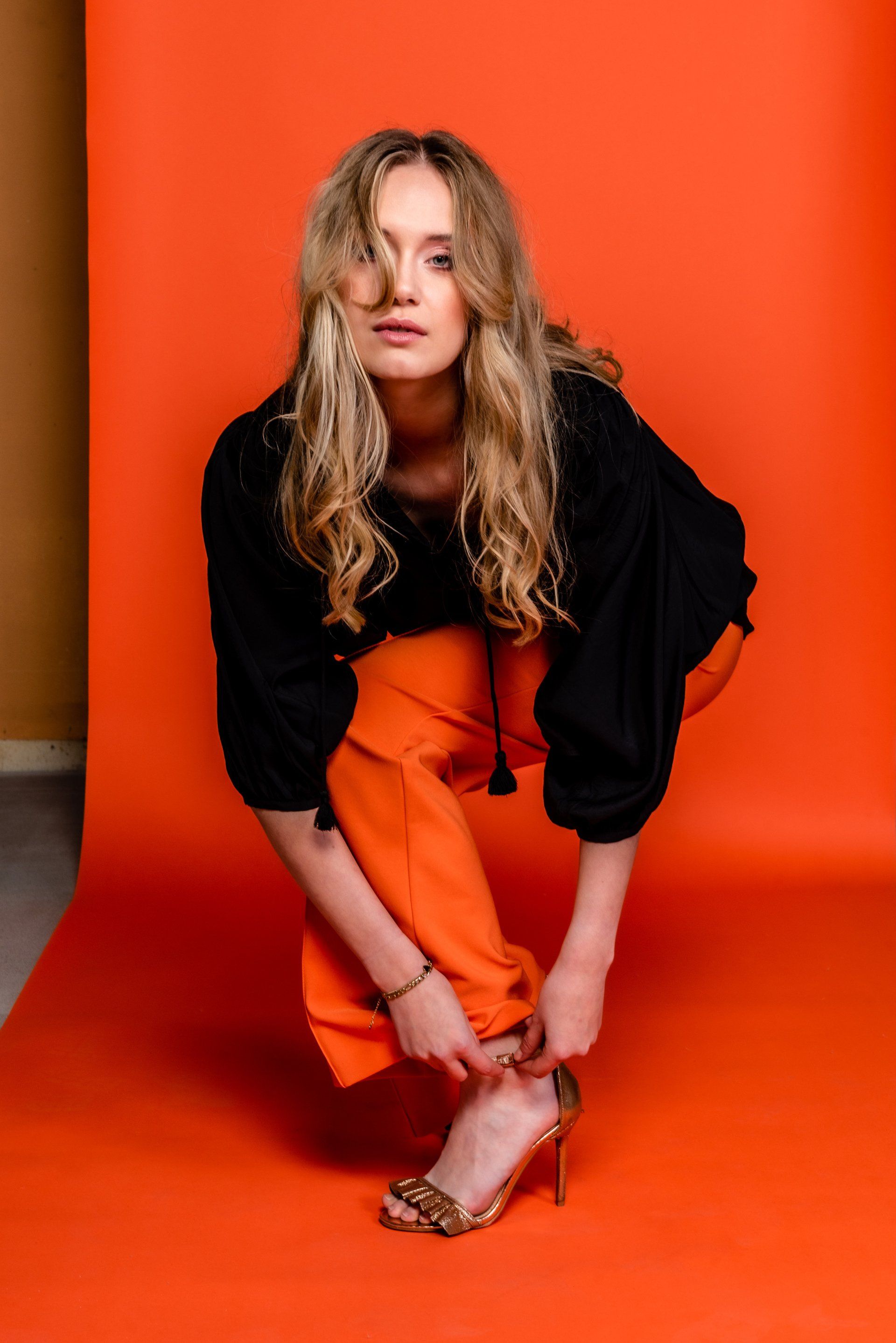 LoisVrouw hurkt op een oranje achtergrond, gekleed in een zwarte top en een oranje broek.