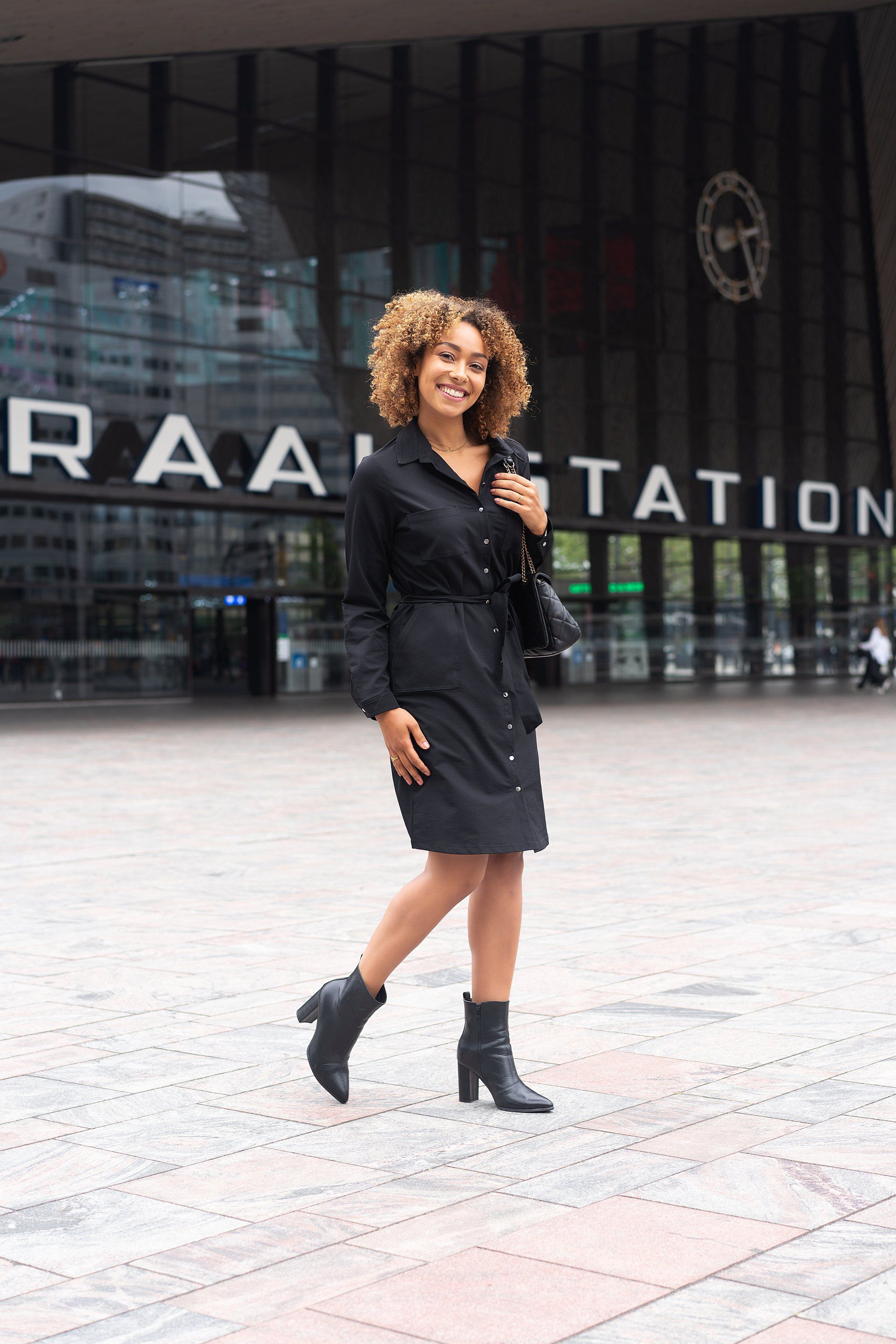 Vrouw in een zwarte jurk loopt buiten een modern glazen gebouw met grote stationsborden.