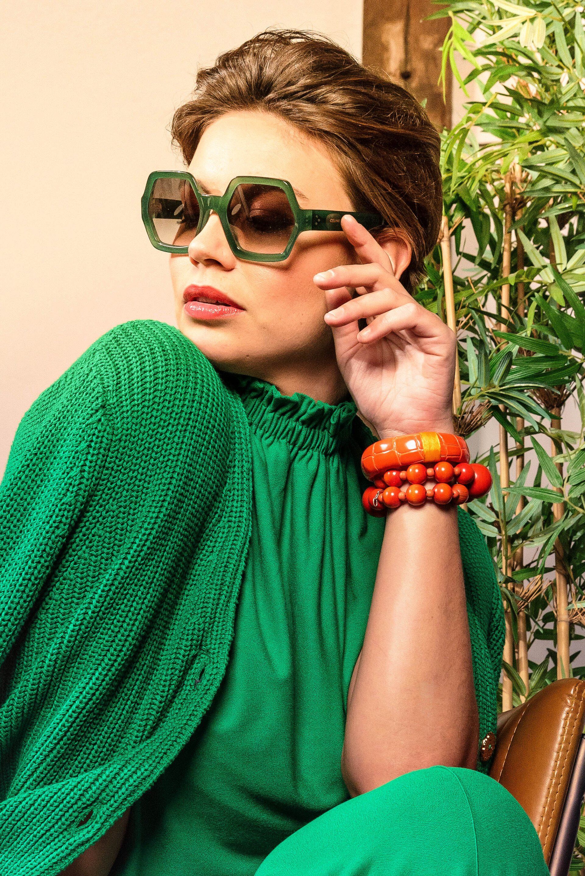 Persoon in een felgroene outfit die een oversized zonnebril rechtzet en oranje armbanden draagt, is buiten.