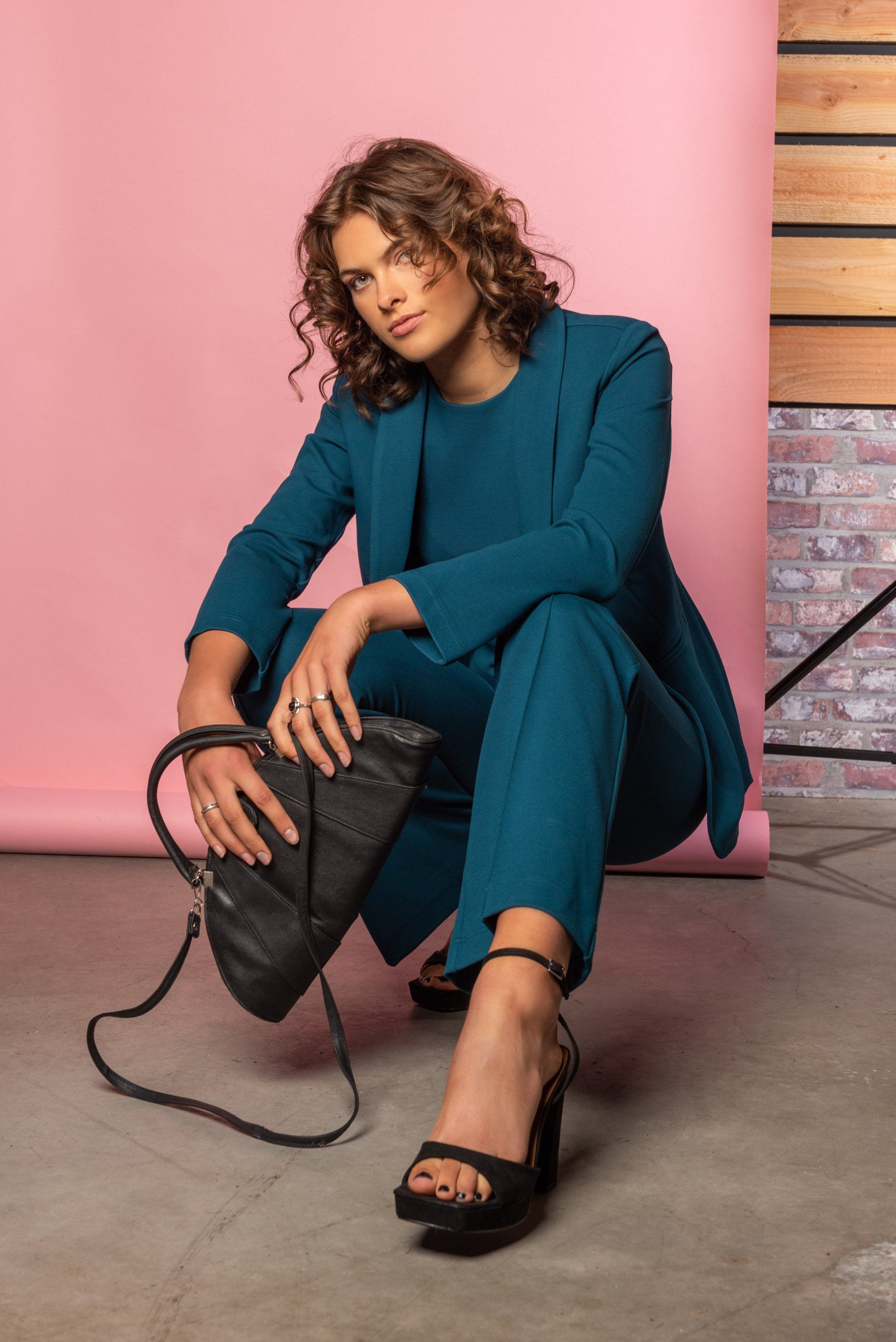 Vrouw in een turquoise pak zit op de grond met een zwarte tas tegen een roze muur.