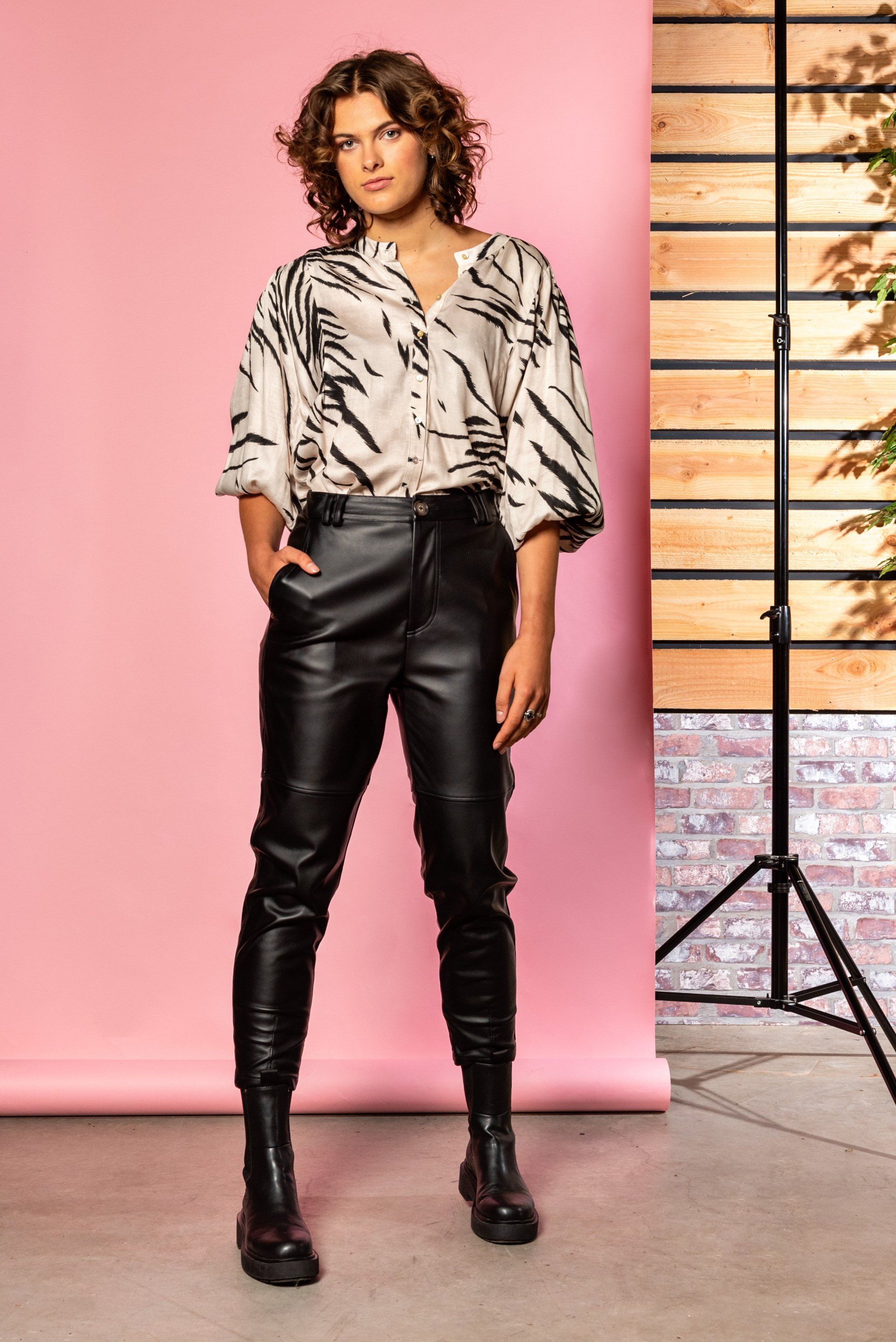 Vrouw in zwarte leren broek en blouse met patroon staat voor een roze achtergrond met een studiolampstatief.