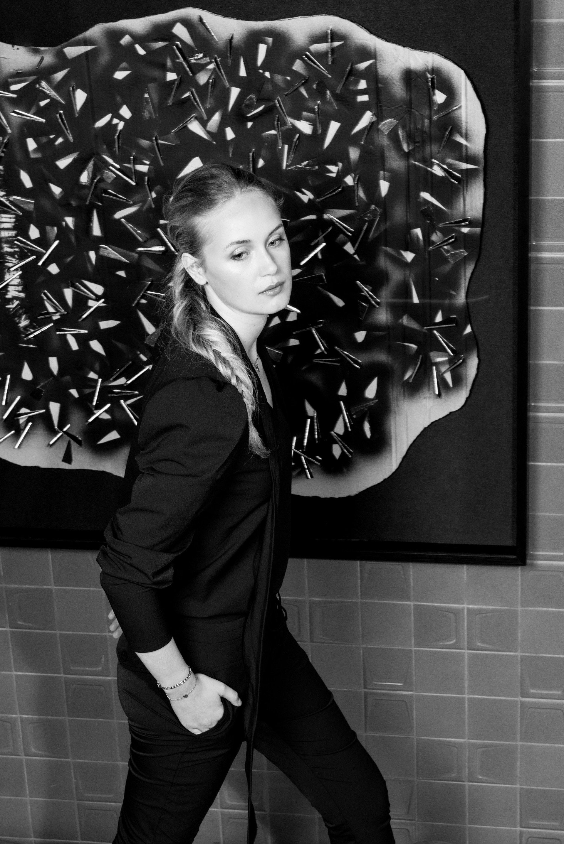 Lois in zwarte kleding poseert voor een groot abstract kunstwerk op een betegelde muur.