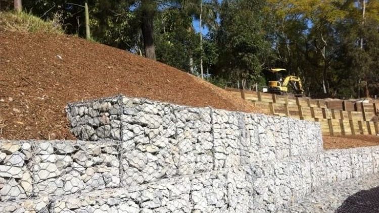 Stone retaining wall beside a graded hillside, with a small excavator in the distance.