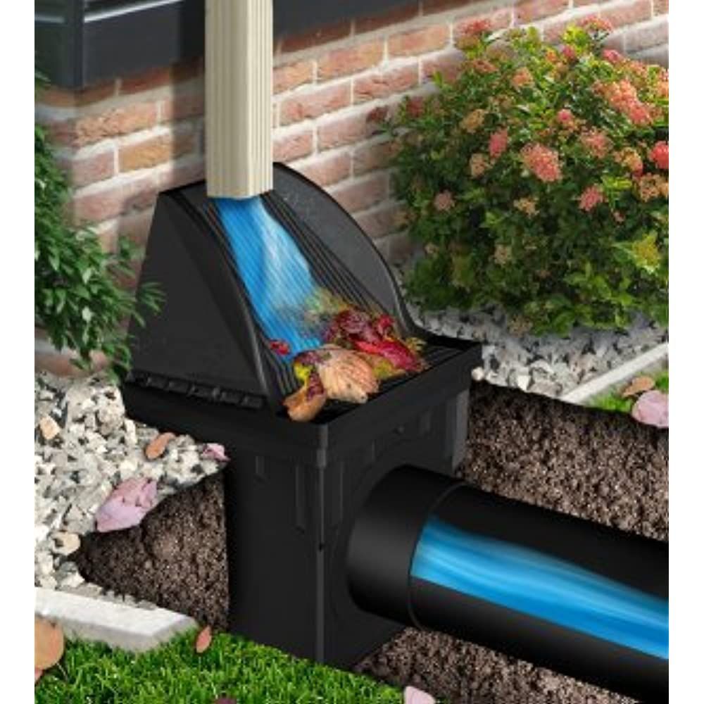 Outdoor drain box with blue water flowing from a downspout into a buried pipe beside a brick wall and shrubs