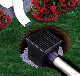 Black yard drain box with grate, surrounded by grass, gravel, flowers, and a white pipe.
