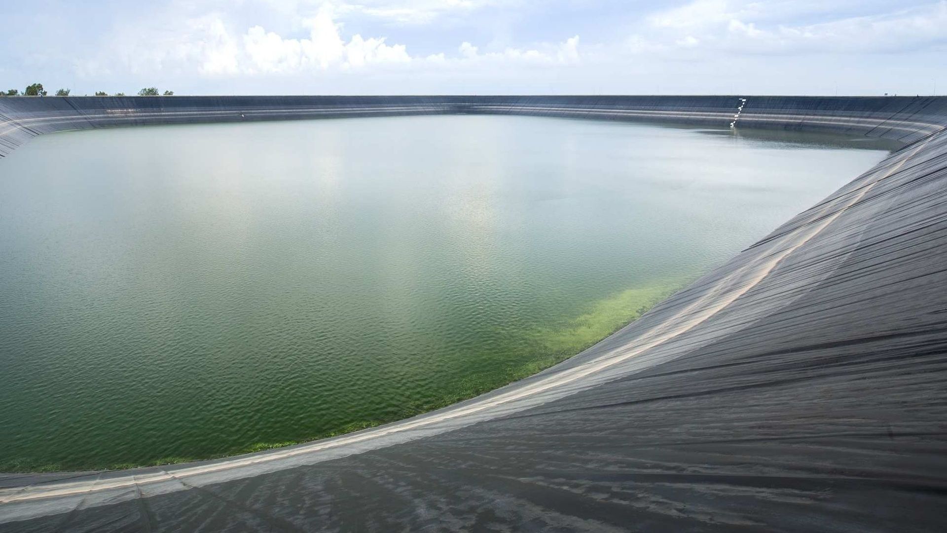 Curved concrete dam wall bordering a green reservoir under a cloudy sky
