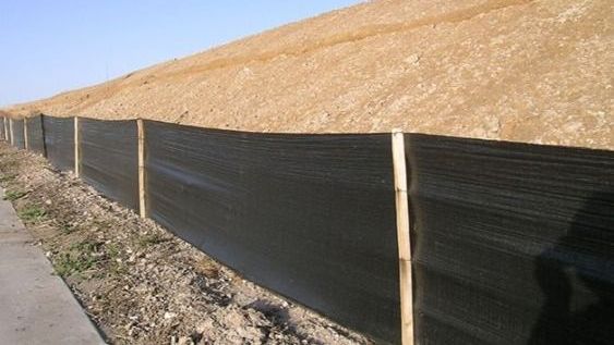 Black mesh fence along a sidewalk beside a dry, grassy hillside under a clear sky