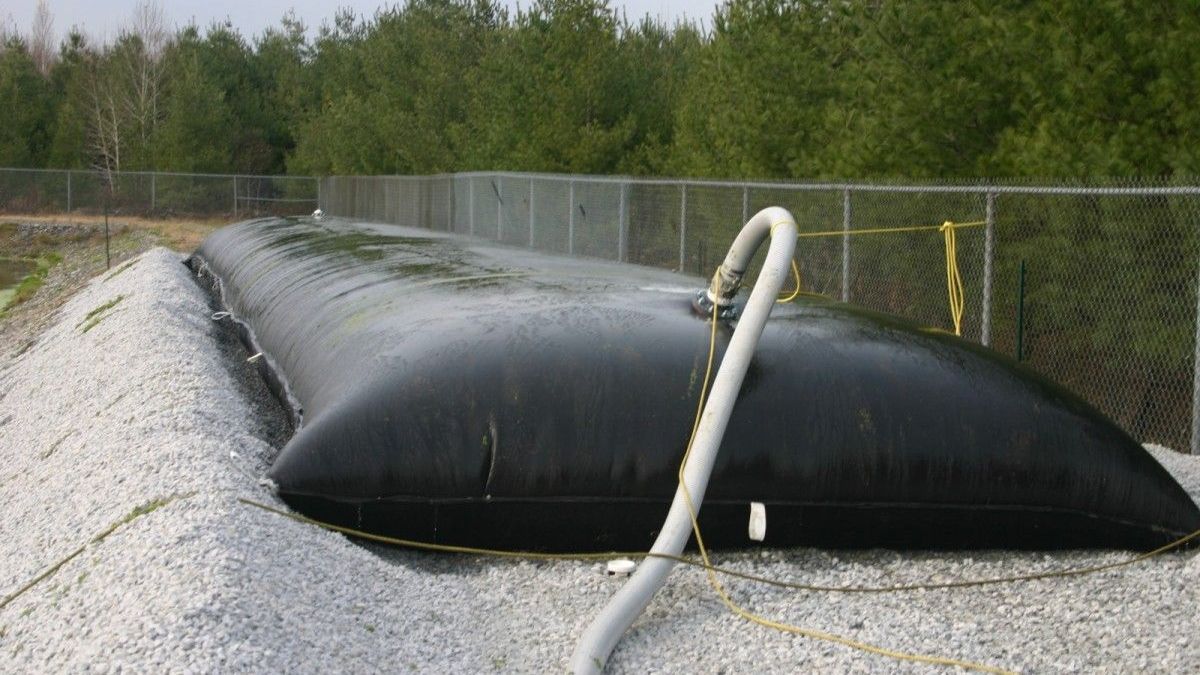 Black inflatable barrier along gravel shore by chain-link fence, with hose attached, beside water.