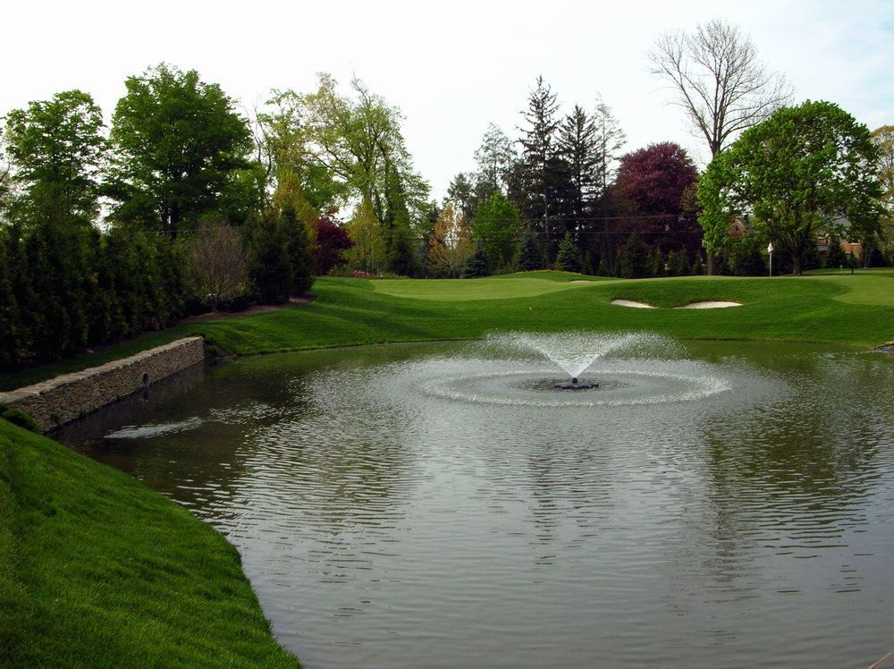 Landscape of a golf course pond with a fountain, grassy banks, and trees under a cloudy sky
