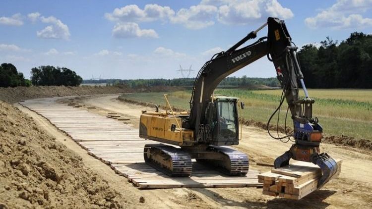 Excavator lifting a bundled load on a construction site beside a dirt road under a blue sky