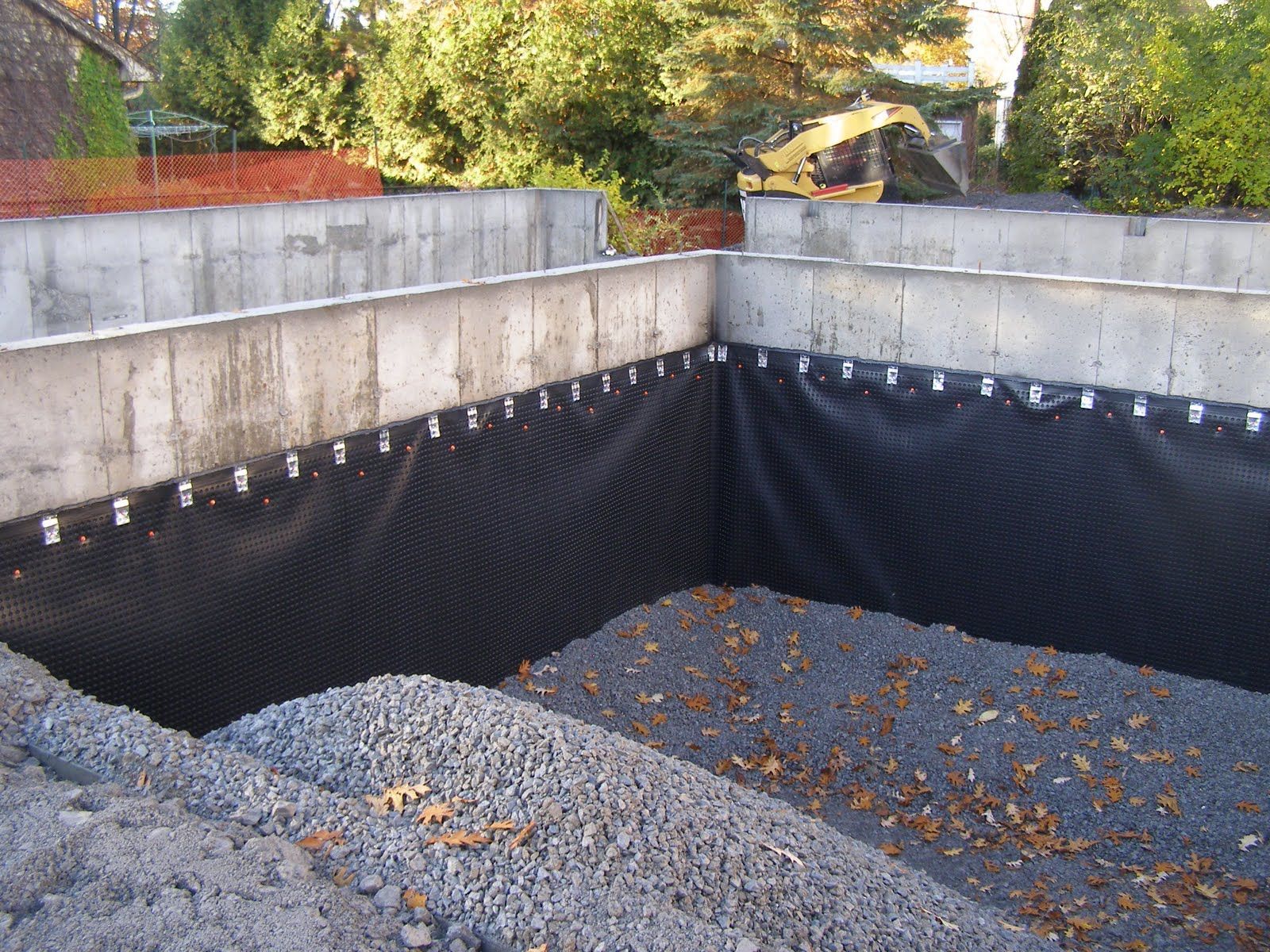 Concrete retaining wall lined with black waterproofing, with gravel and fallen leaves inside a backyard pit.