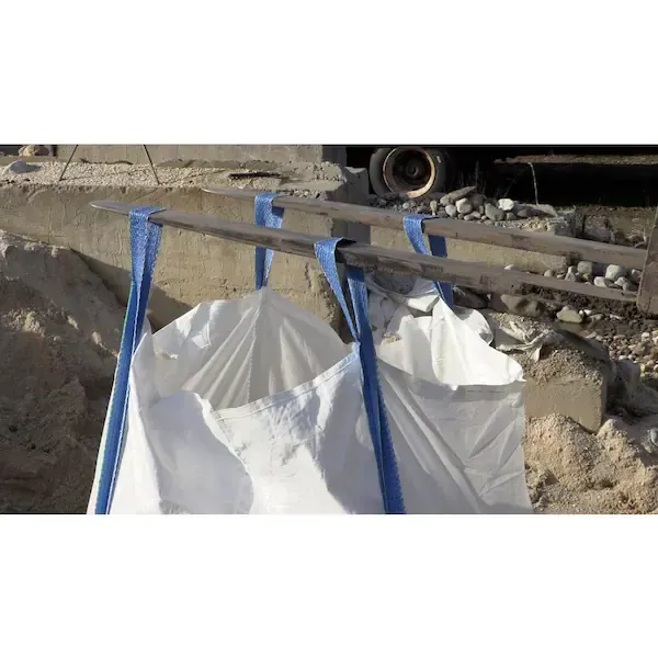 Blue straps hold a large white bulk bag at a construction site near debris and concrete barriers