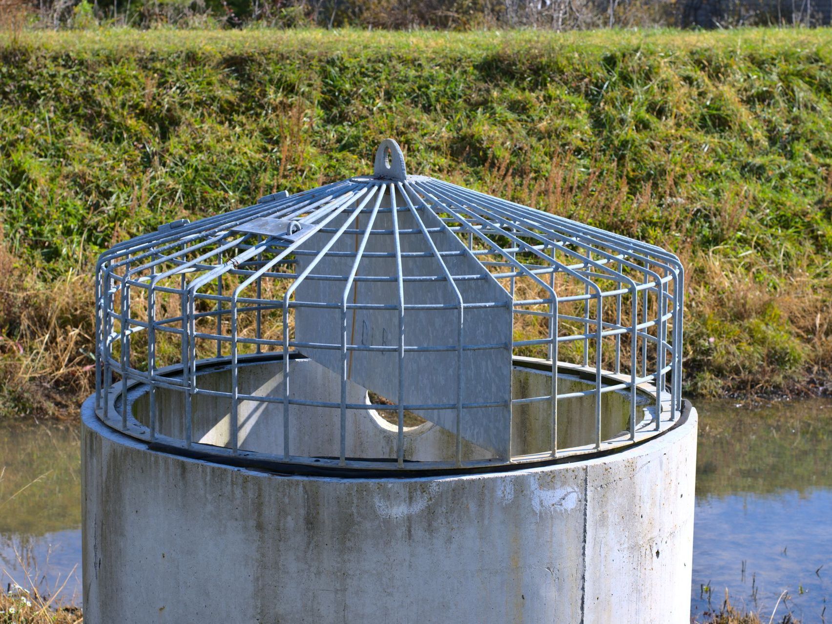 Metal cage over a concrete well beside a water channel and grassy embankment