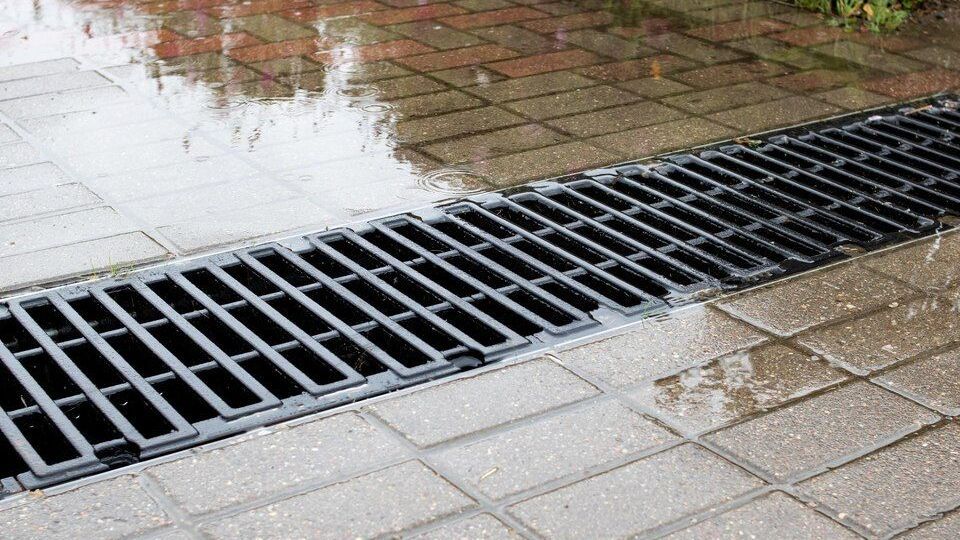 Wet sidewalk with a long black metal storm drain grate along the curb.