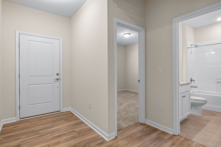 Empty hallway with beige walls, wood flooring, a white door, and a bathroom entrance on the right