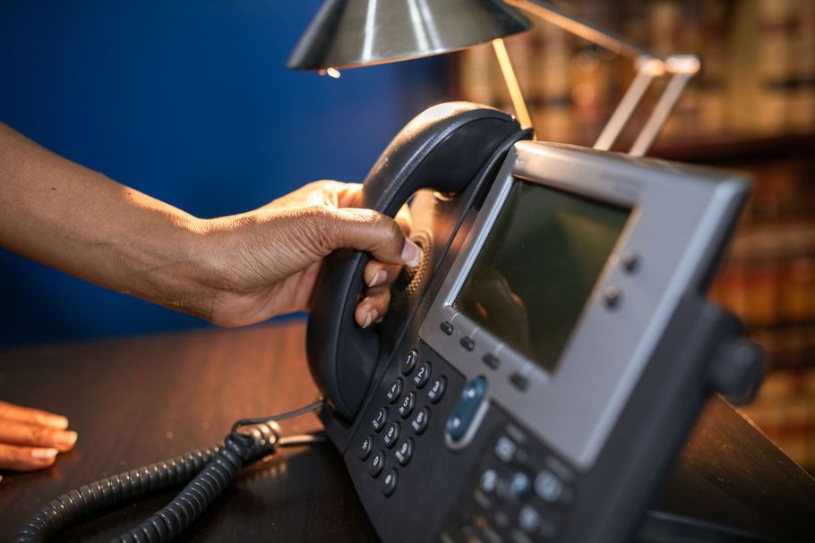 Hand holding a black office phone handset beside a desk lamp and keypad on a desk